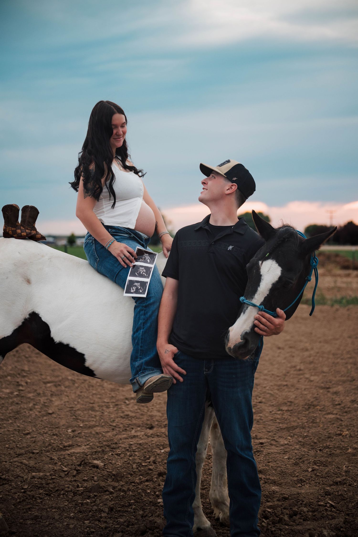 Pregnant woman sitting on a black and white horse holding ultrasound photos while man in cowboy hat stands beside her.