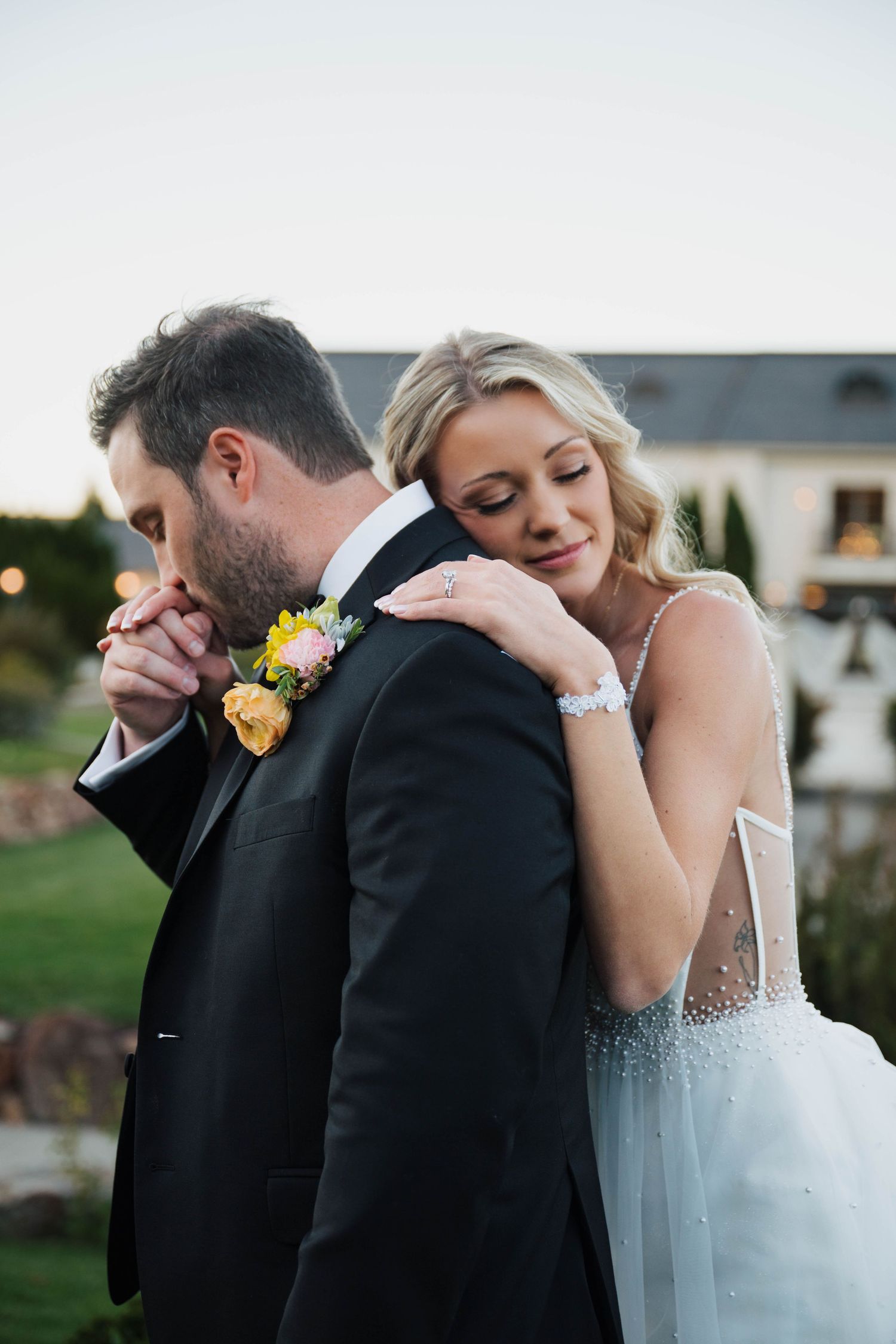 Bride rests her head on groom's shoulder in a tender wedding moment, both dressed elegantly outdoors at dusk.