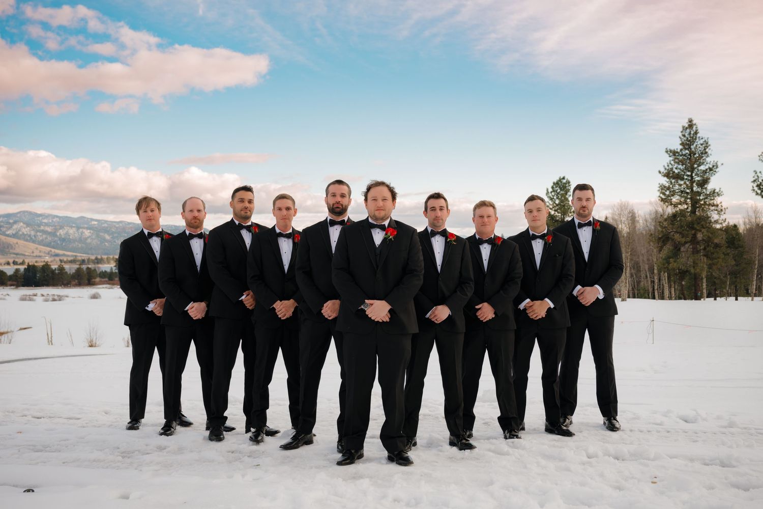 Groomsmen in black tuxedos pose together in a snowy winter landscape with mountains in the background.