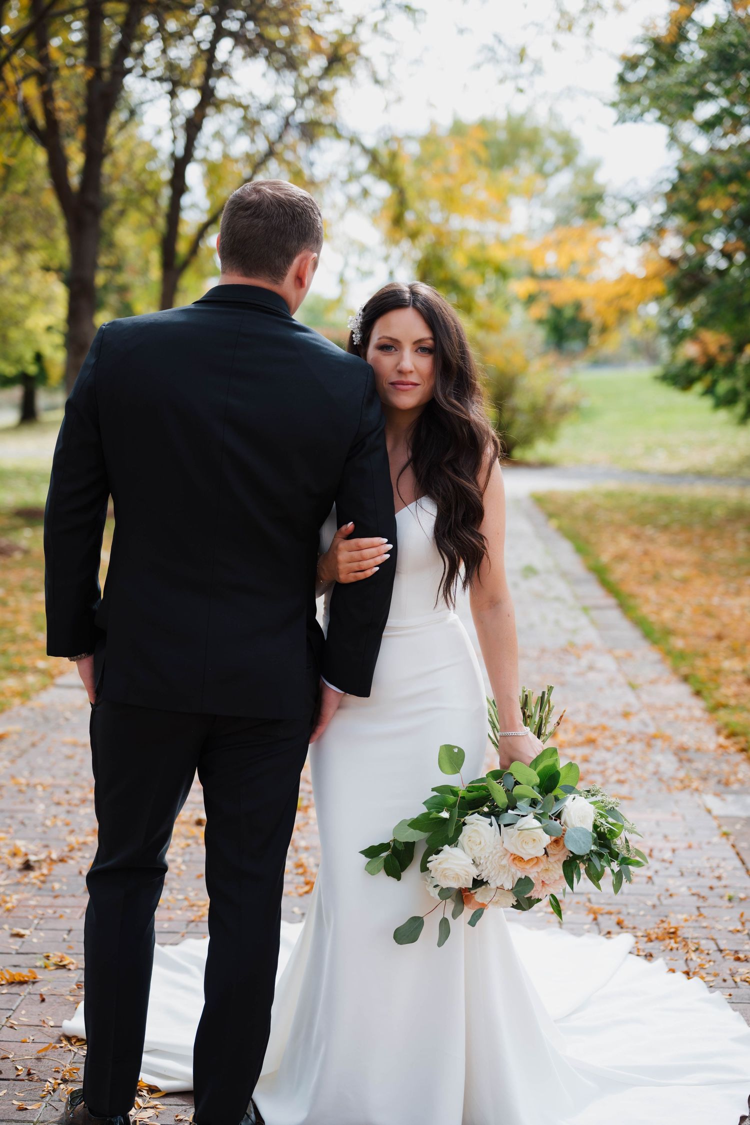 Bride in white gown holds bouquet while walking with groom in black suit on autumn park path.