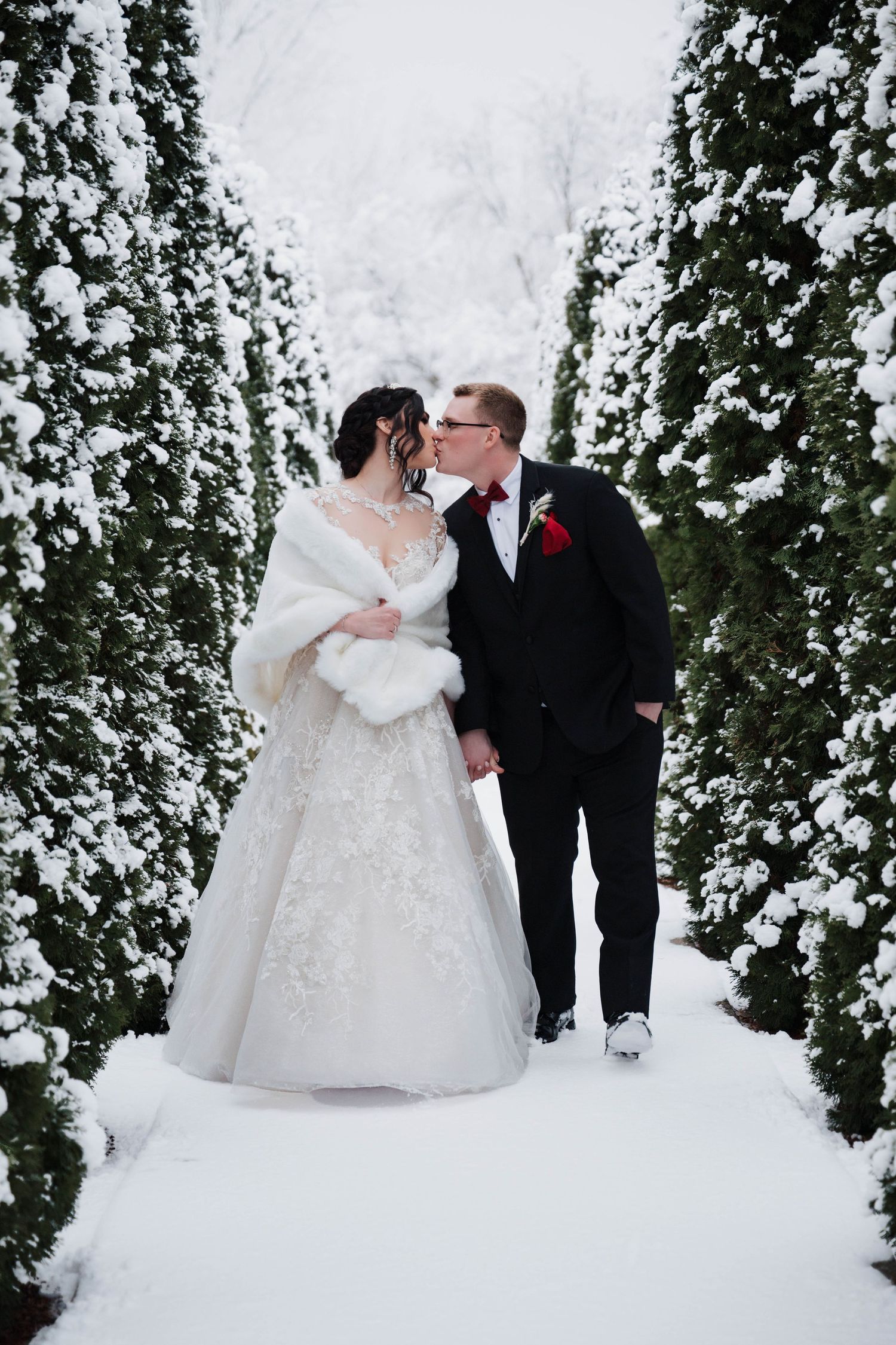 Bride and groom kissing in snowy winter garden path lined with snow-covered evergreen trees on wedding day.