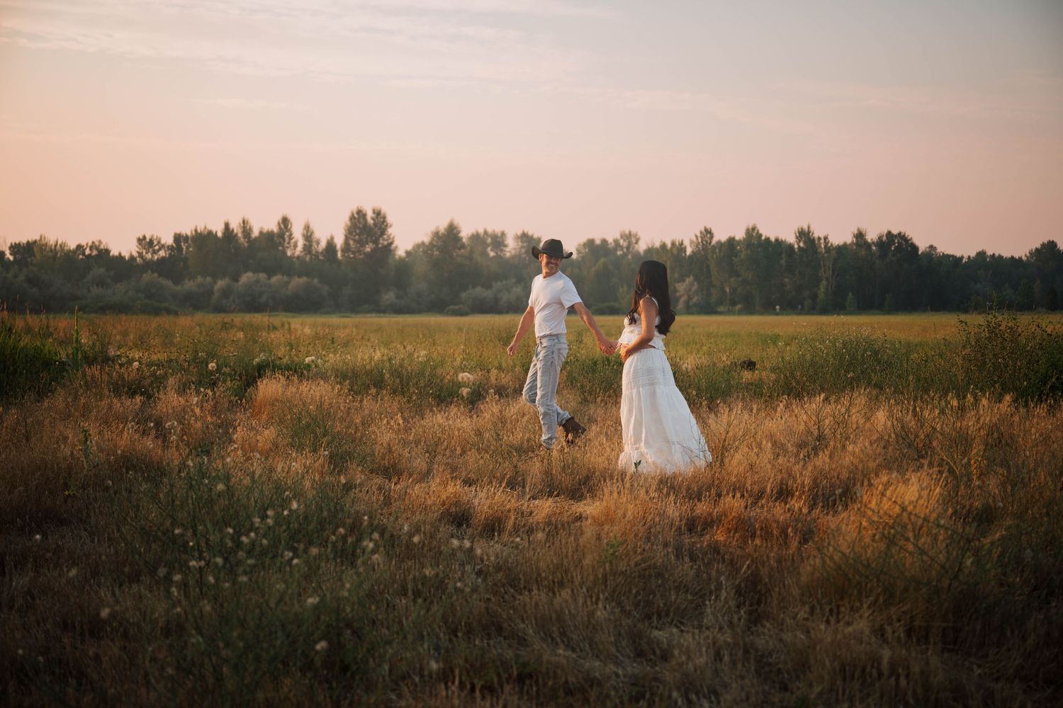 Couple in white attire walking through golden field at sunset with trees in background.