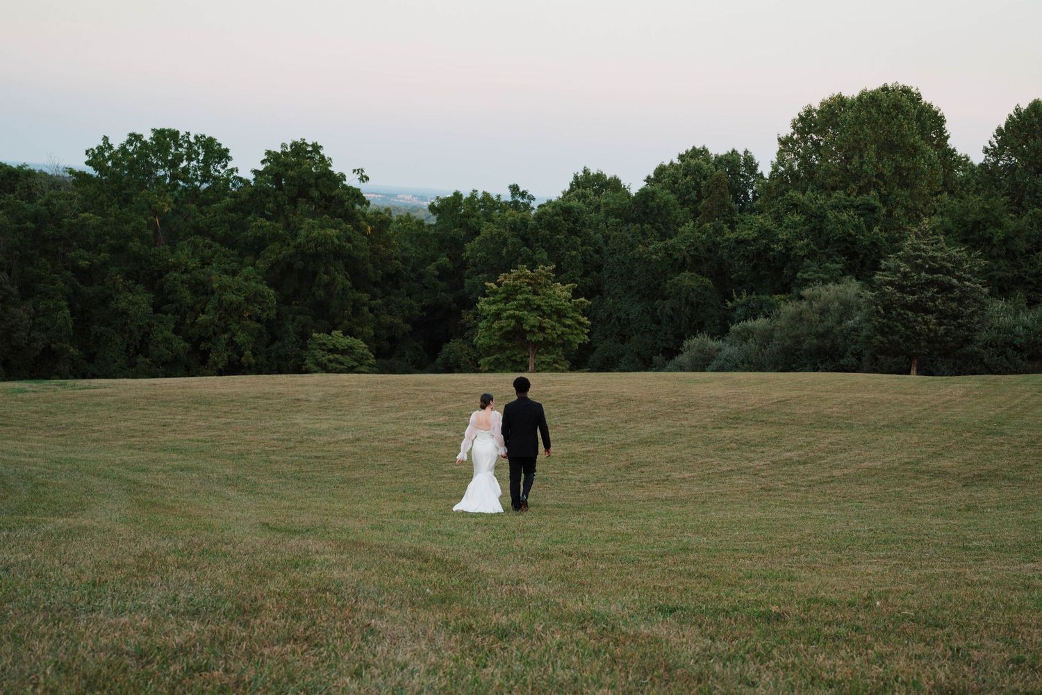 Bride and groom walking hand in hand across a lush green field at dusk with trees in the background.