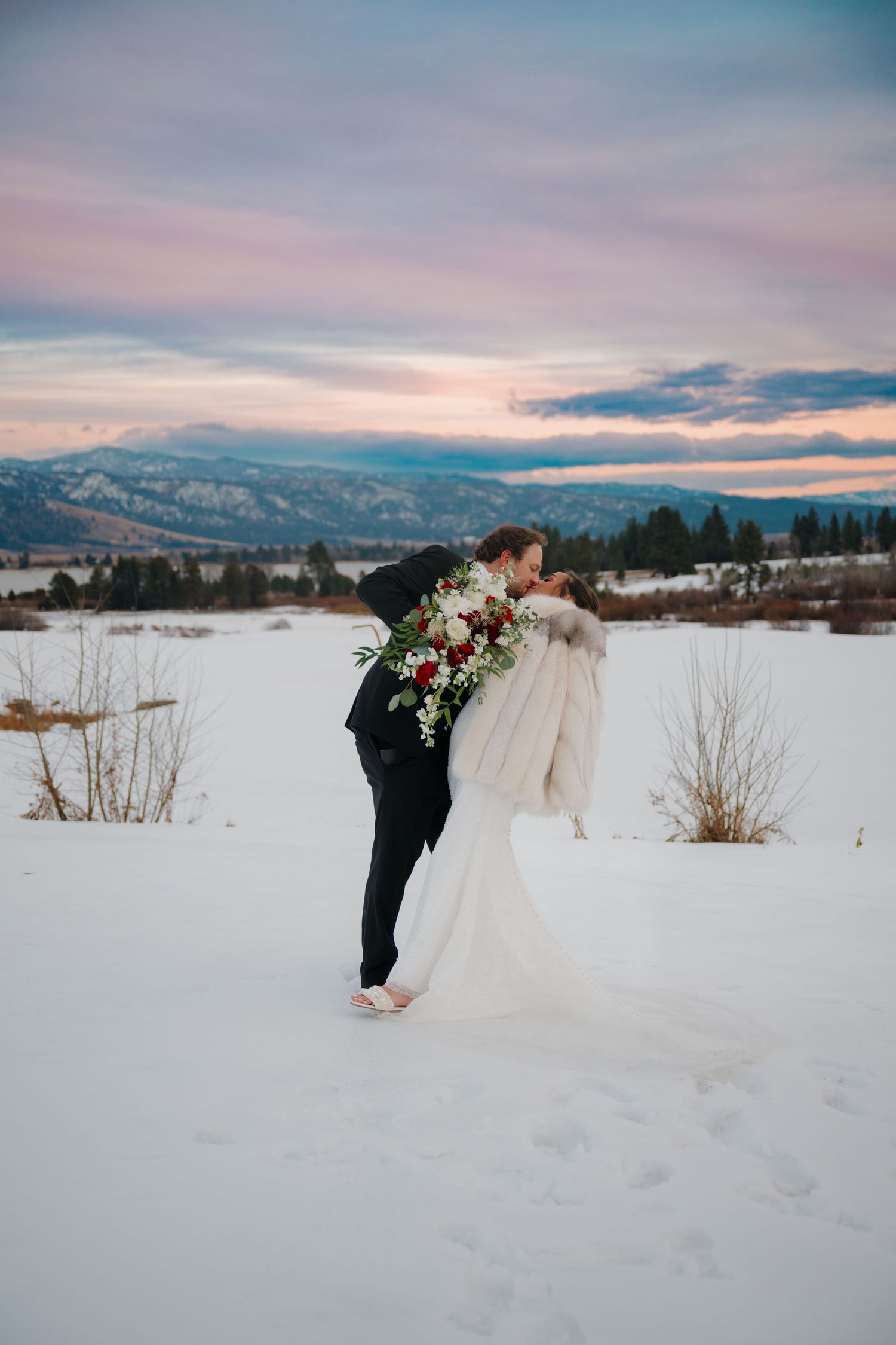 A couple shares a romantic moment in a snowy field during sunset with mountains in the distance.
