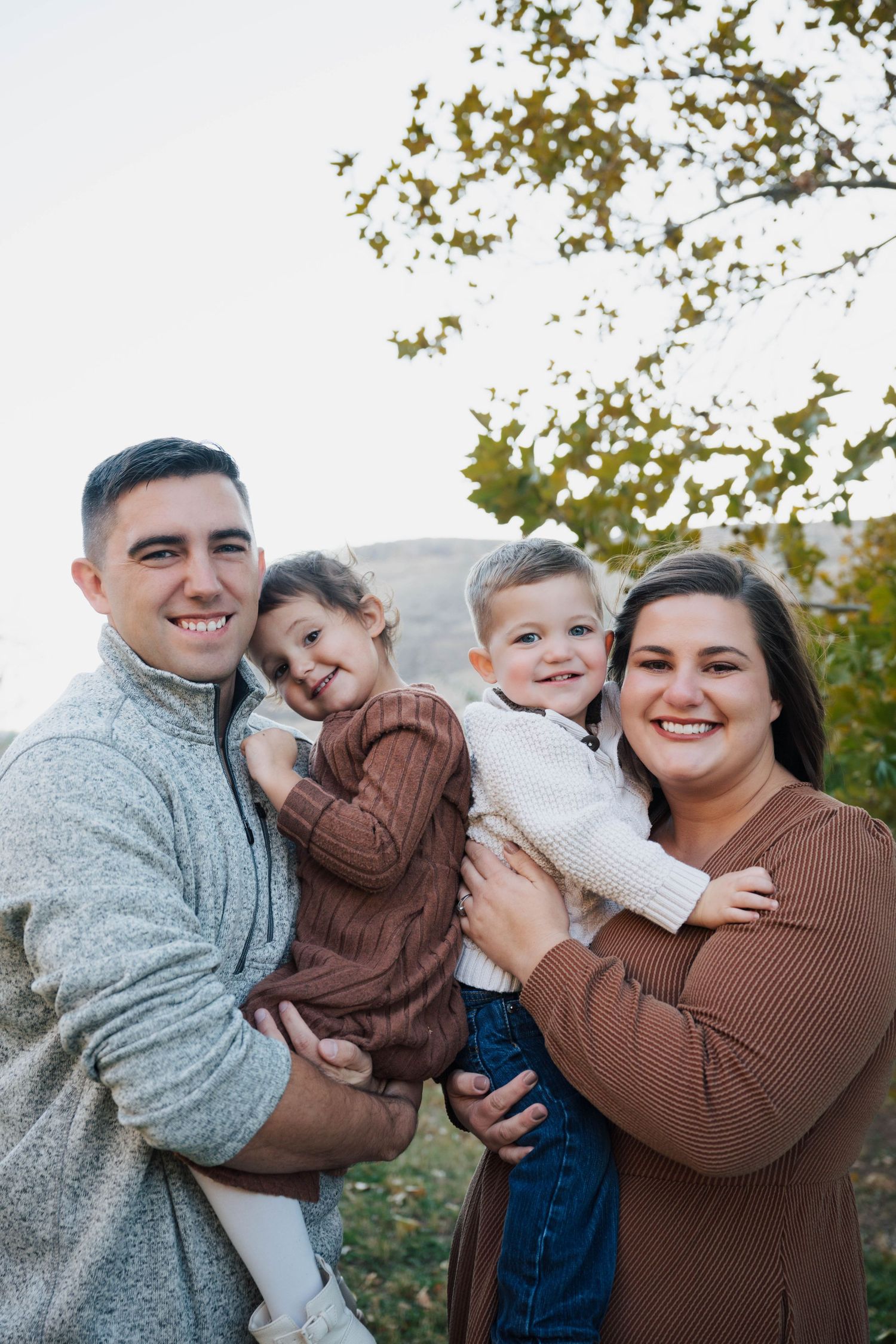Family embraces and laughs together during outdoor photo session wearing coordinated brown and grey outfits.