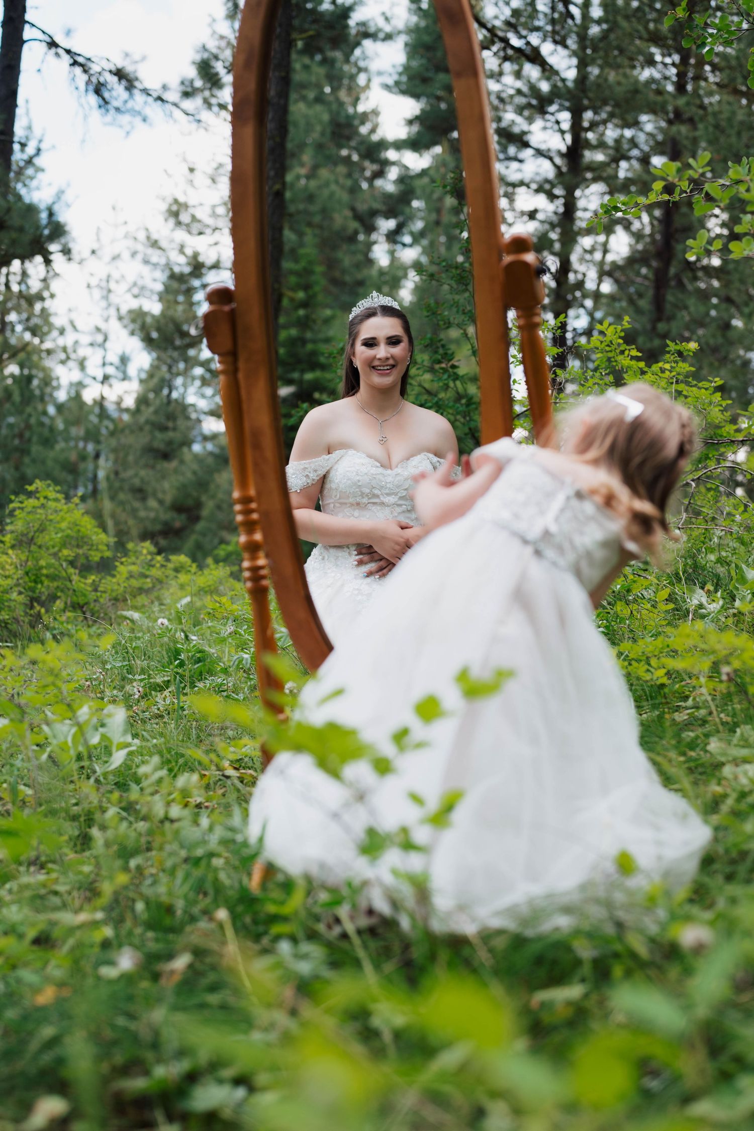 Bride in white gown views her reflection in a wooden mirror outdoors in a lush green forest setting.