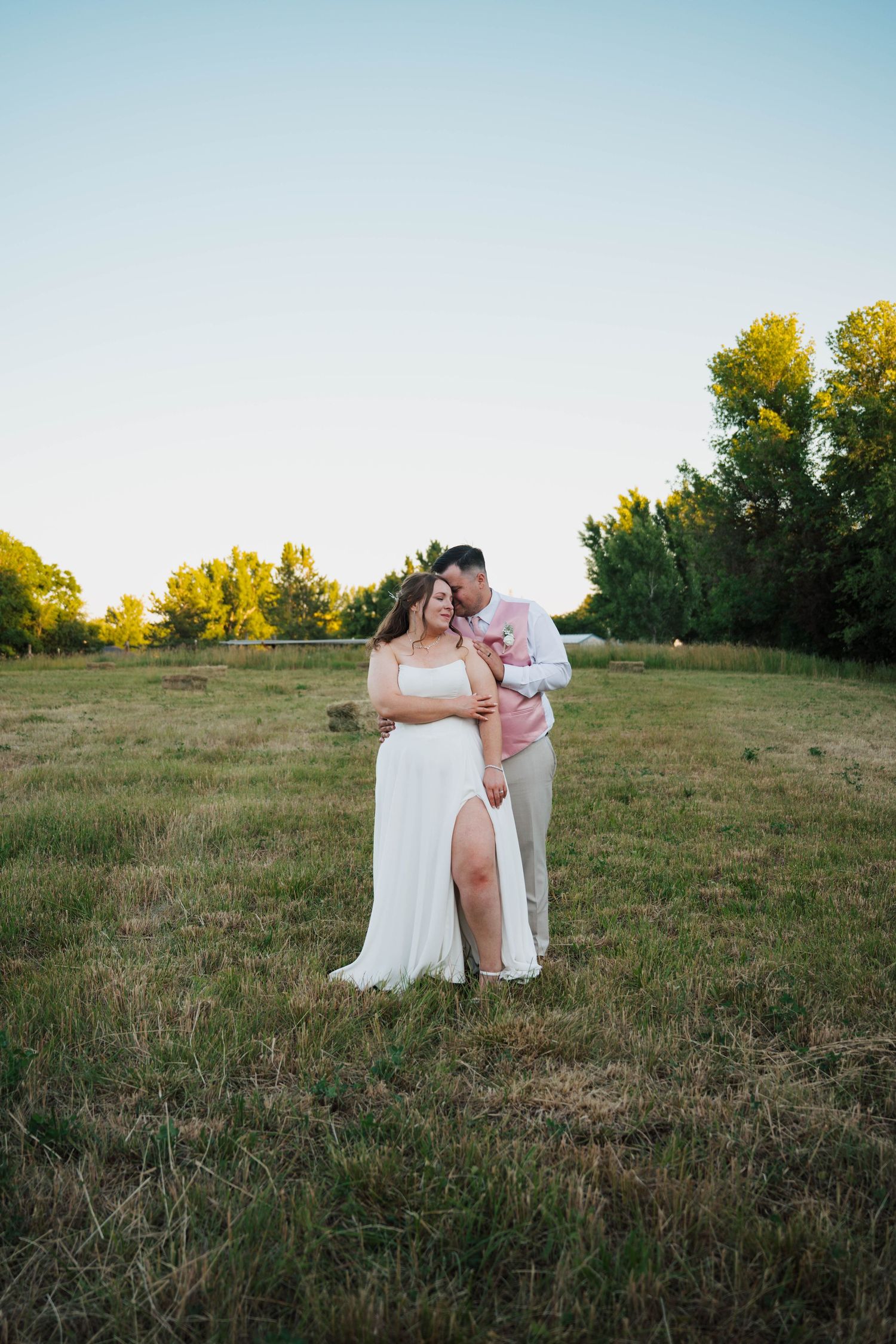 Couple in wedding attire embracing in a green field at golden hour, bride in white gown, groom in pink suit.