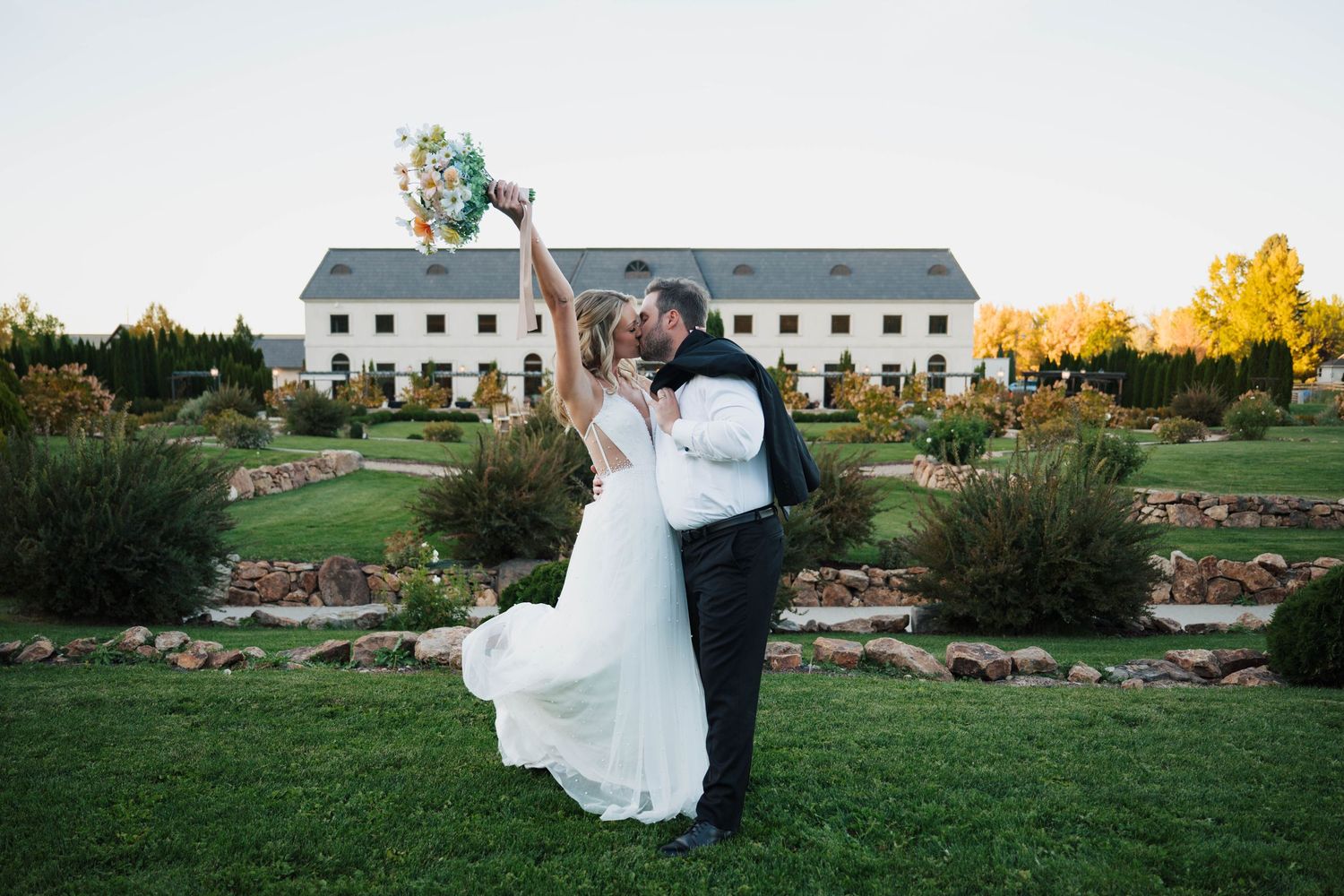 Bride and groom kissing on lush green lawn with bouquet raised, elegant white venue building in background at golden hour.