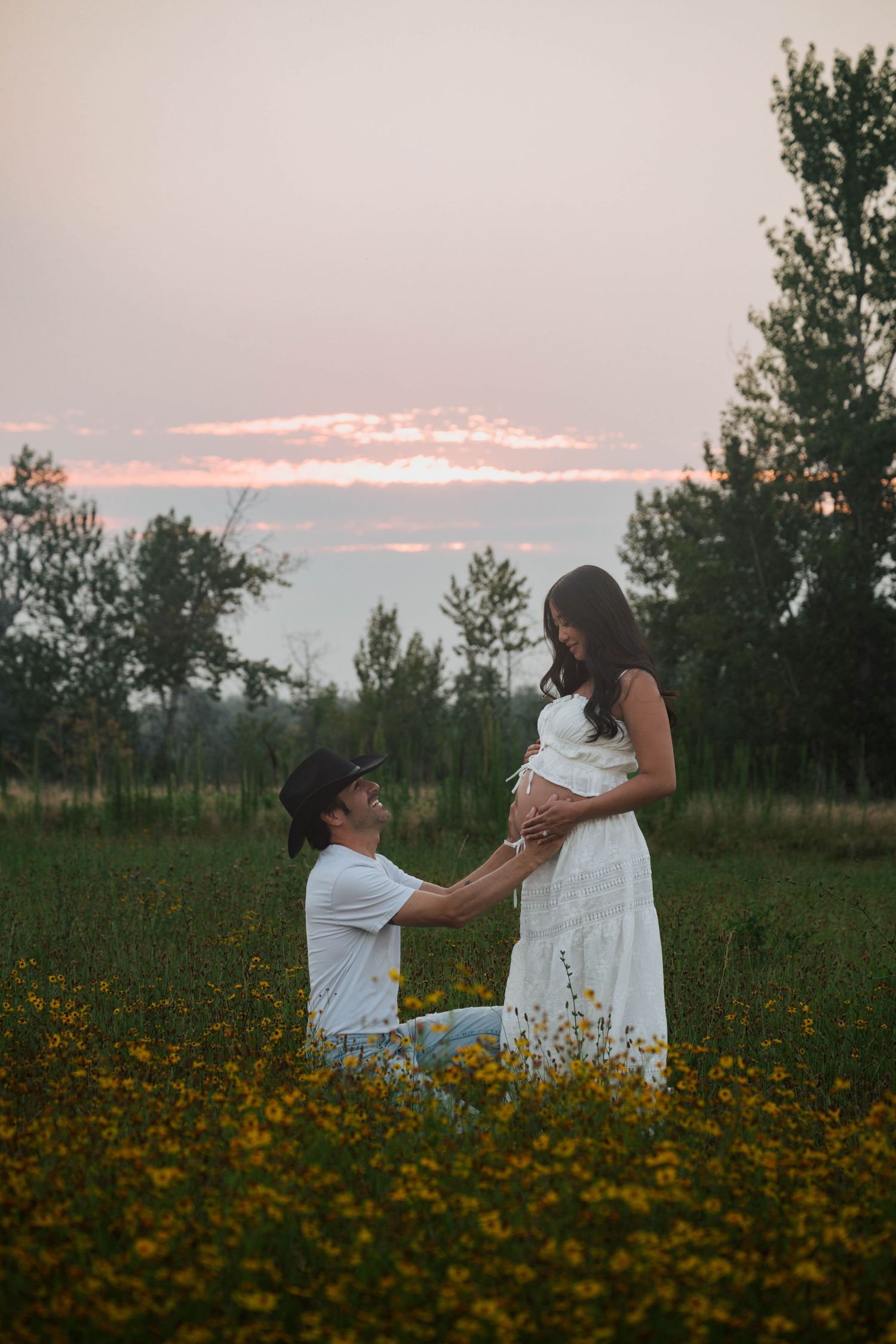 Man kneeling before pregnant woman in white dress in flower field at sunset.