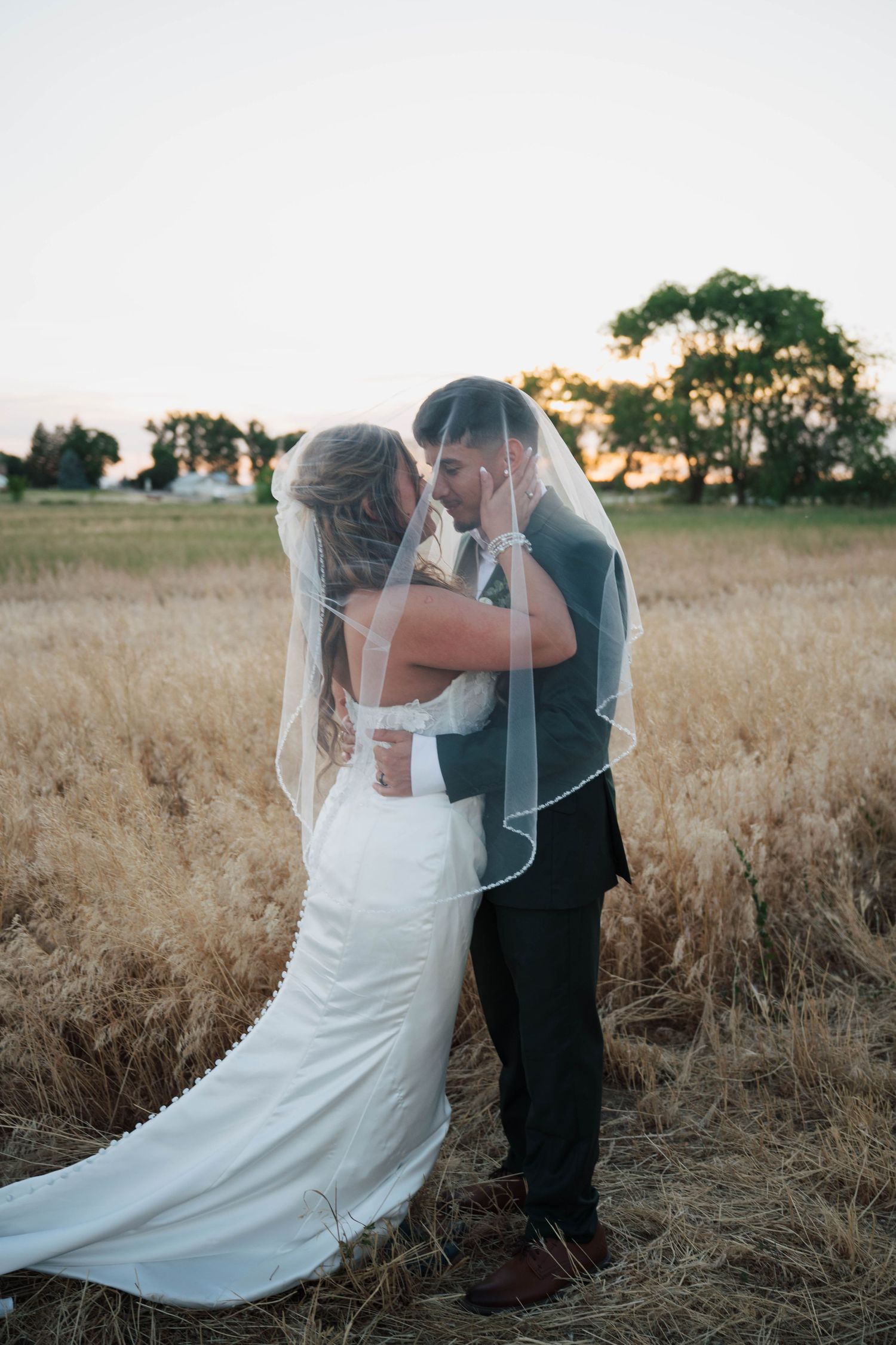 Bride and groom kissing under a veil in a golden field at sunset, surrounded by nature at rustic daisy  event barn in nampa, idaho