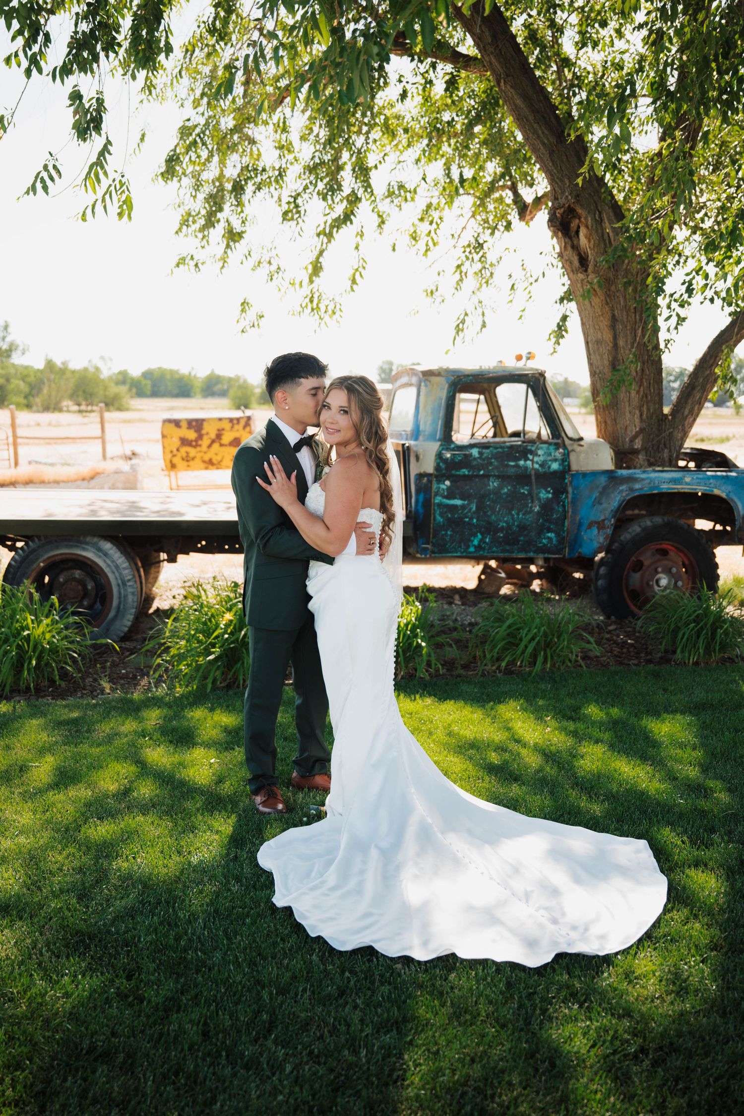Bride and groom embrace on green lawn beside vintage blue truck under large tree on sunny wedding day.