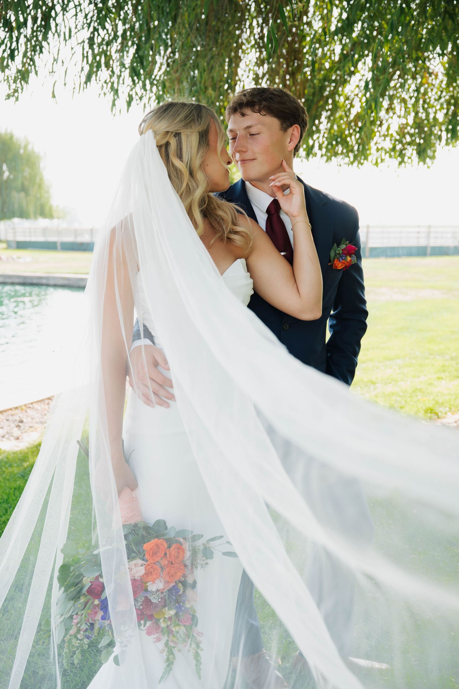 Bride and groom share a romantic moment under a willow tree, her veil flowing, holding a colorful bouquet outdoors.
