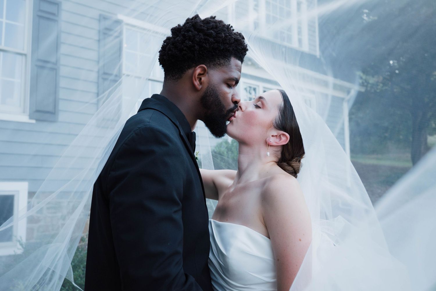 Interracial couple sharing a romantic kiss under a flowing white wedding veil outdoors.