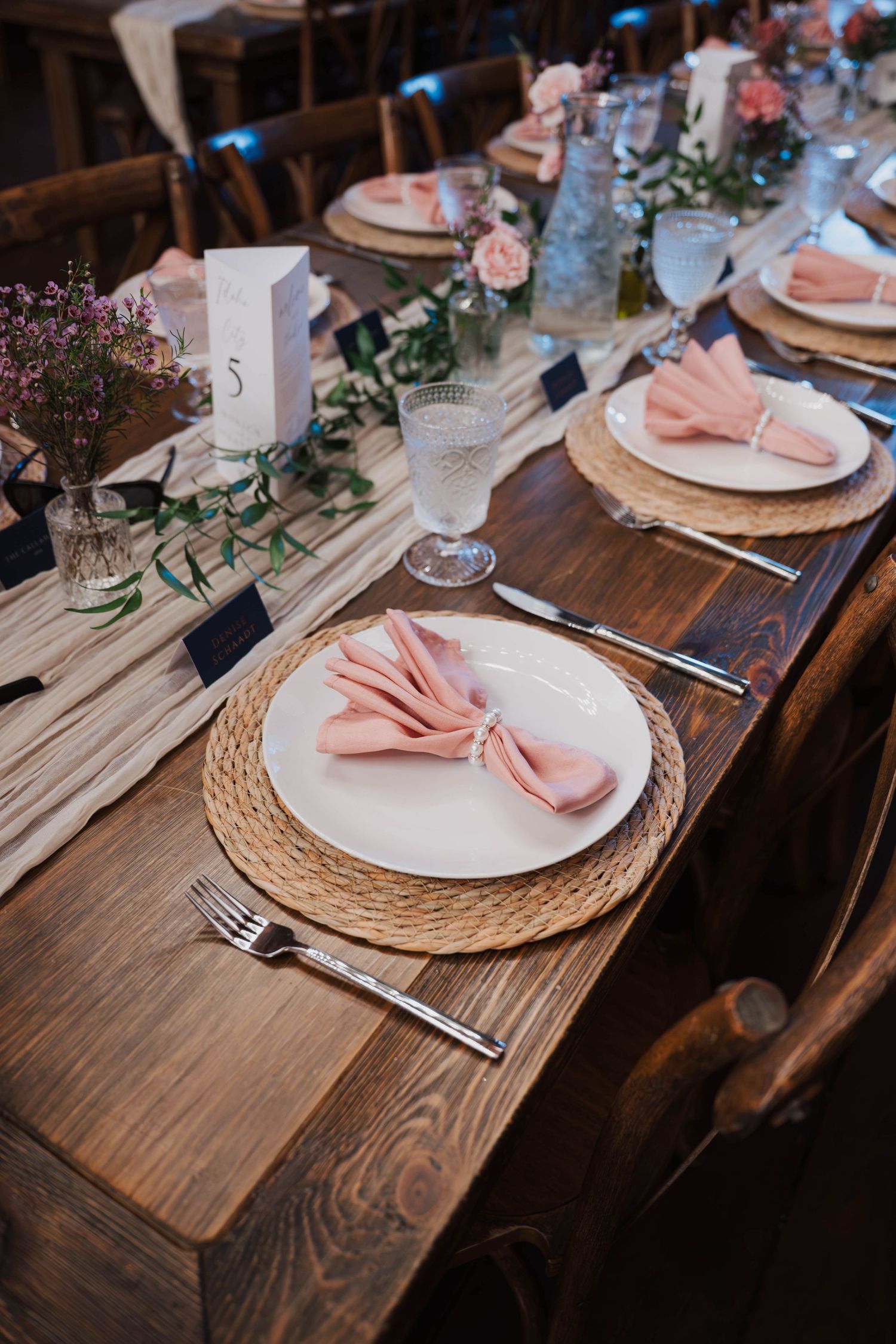 Rustic wedding reception table with pink bow napkins on woven chargers, wildflowers, and greenery on dark wood farmhouse tables.