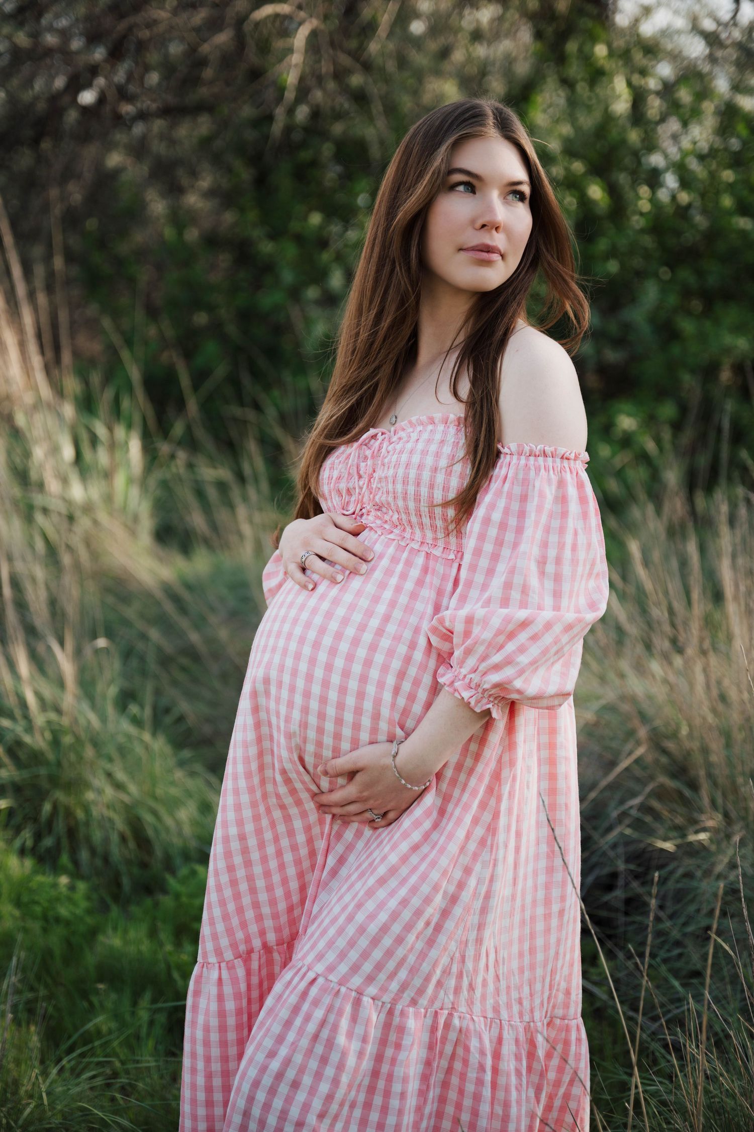 Pregnant woman in pink gingham off-shoulder dress cradling baby bump outdoors in lush green field.