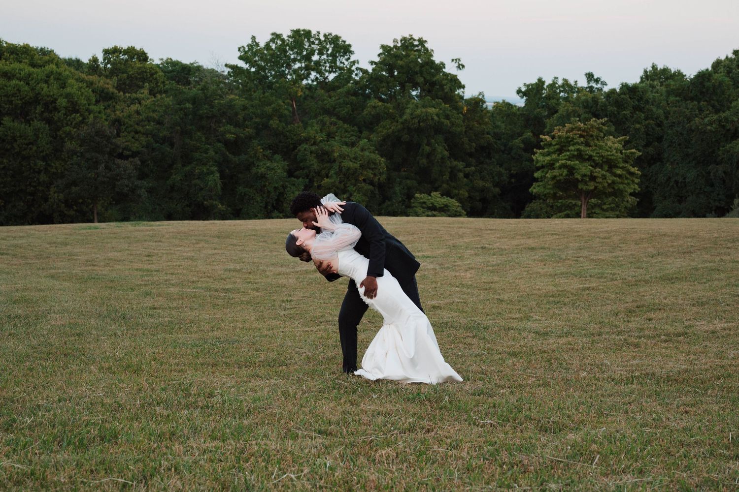 Groom dipping bride in white wedding dress on green field with trees in background.