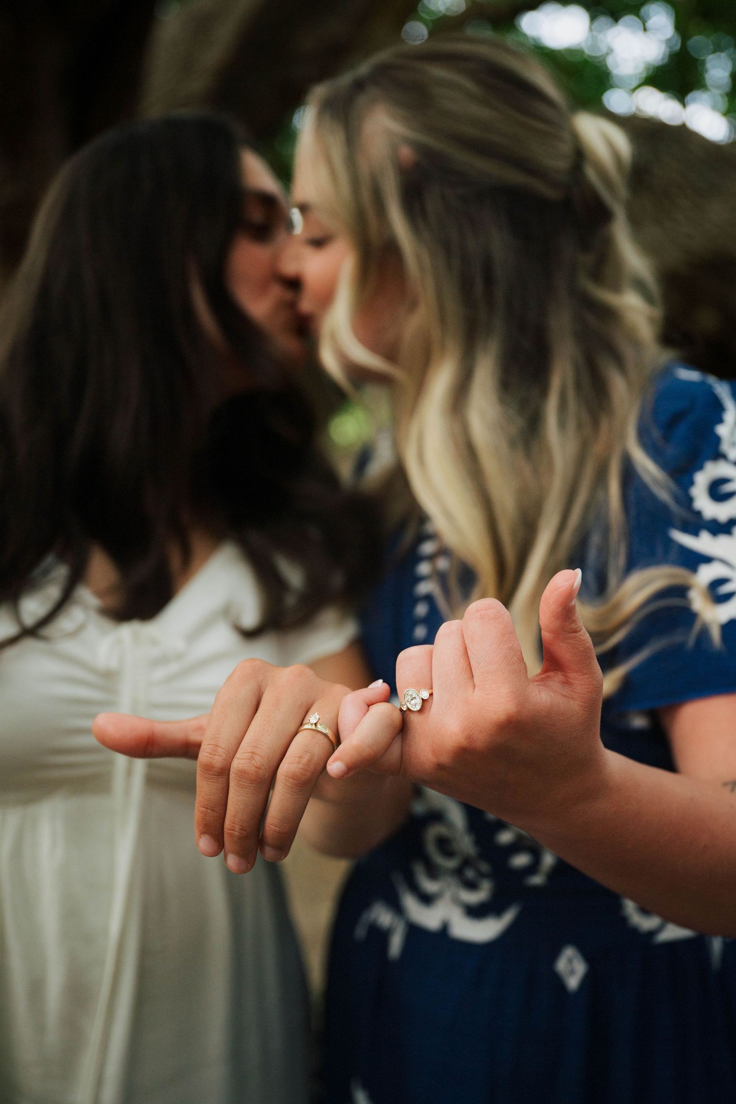 Two women sharing a kiss while displaying engagement rings, one in white dress and one in blue floral dress.