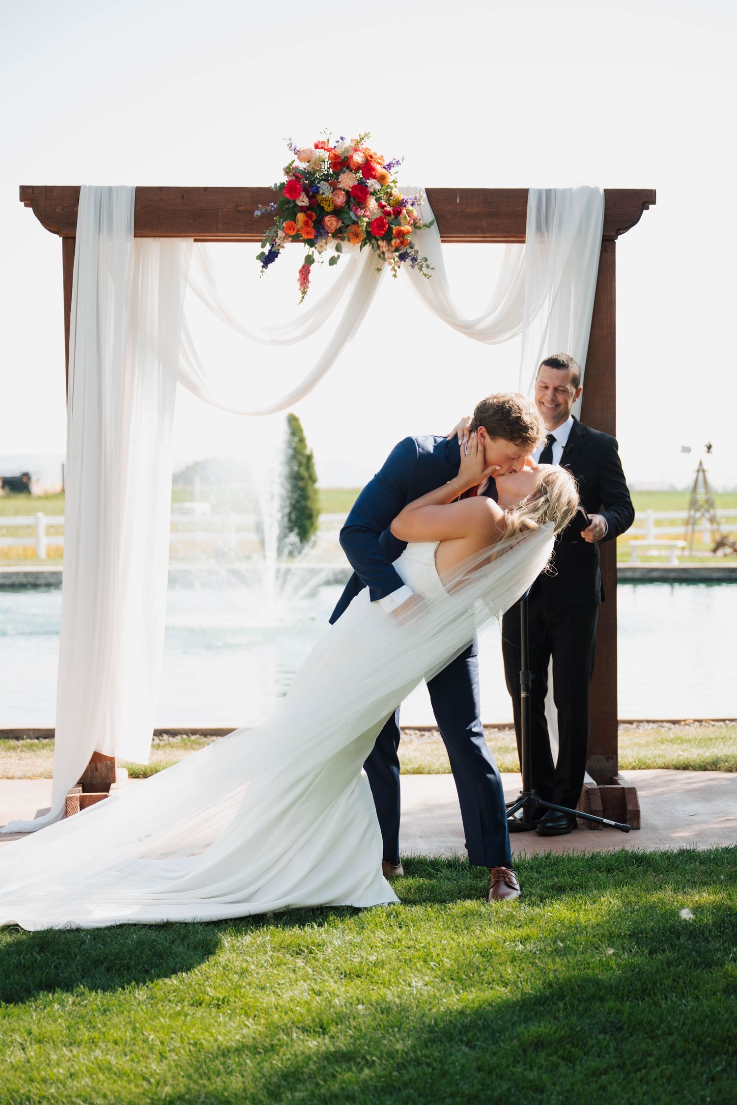 Bride and groom share first kiss dip under floral arch at outdoor wedding ceremony by fountain.