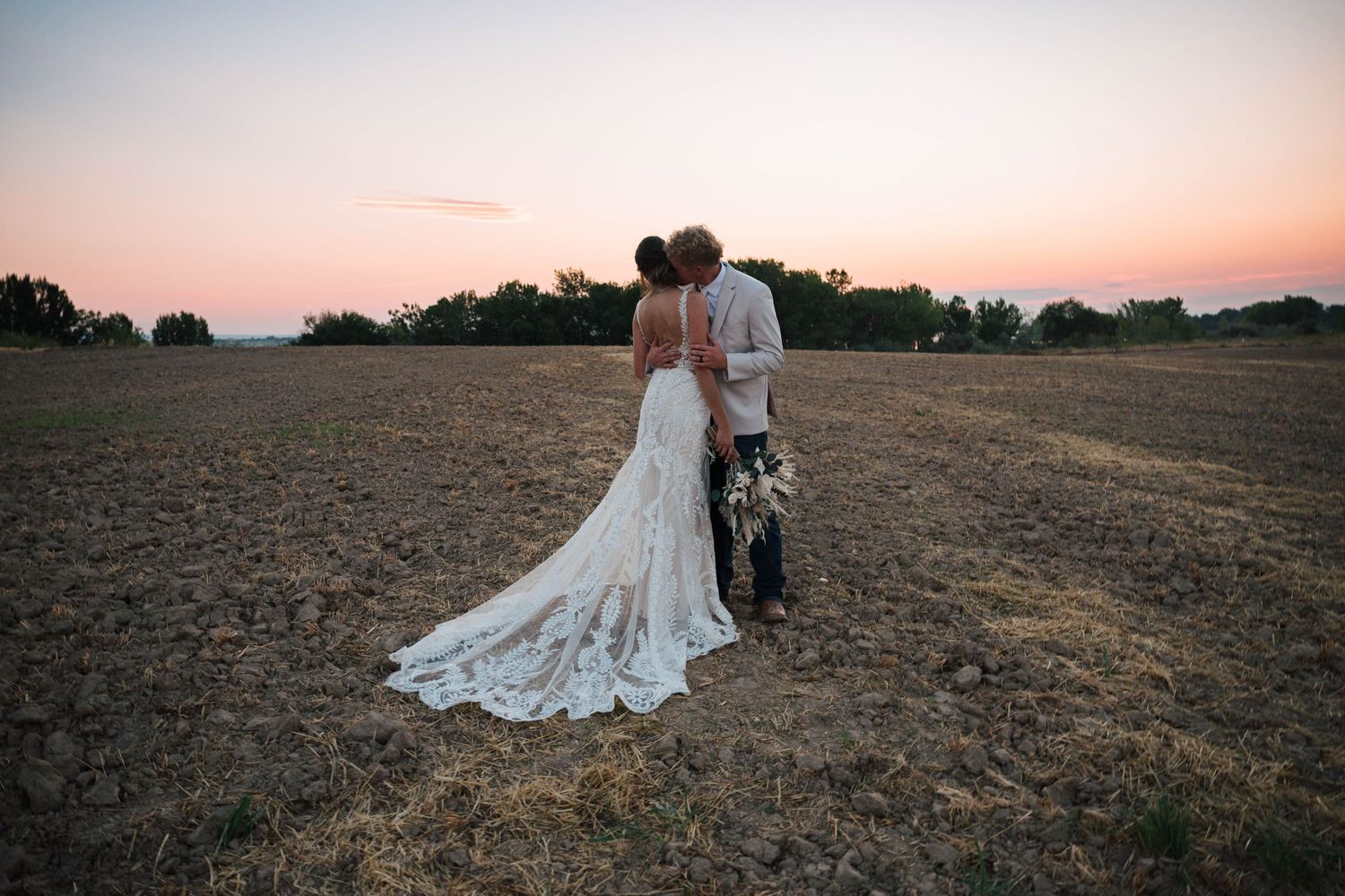 Bride and groom kissing at sunset in an open field, bride wearing a lace wedding gown with a long train.