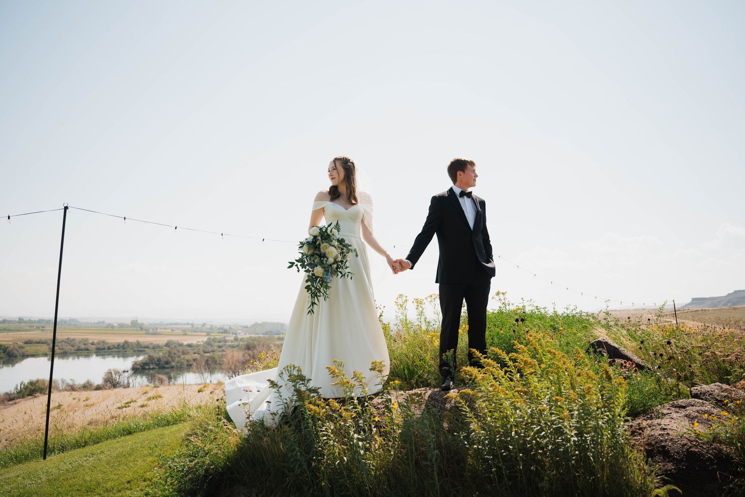 A couple stands on a hilltop overlooking a scenic landscape during their wedding photoshoot.