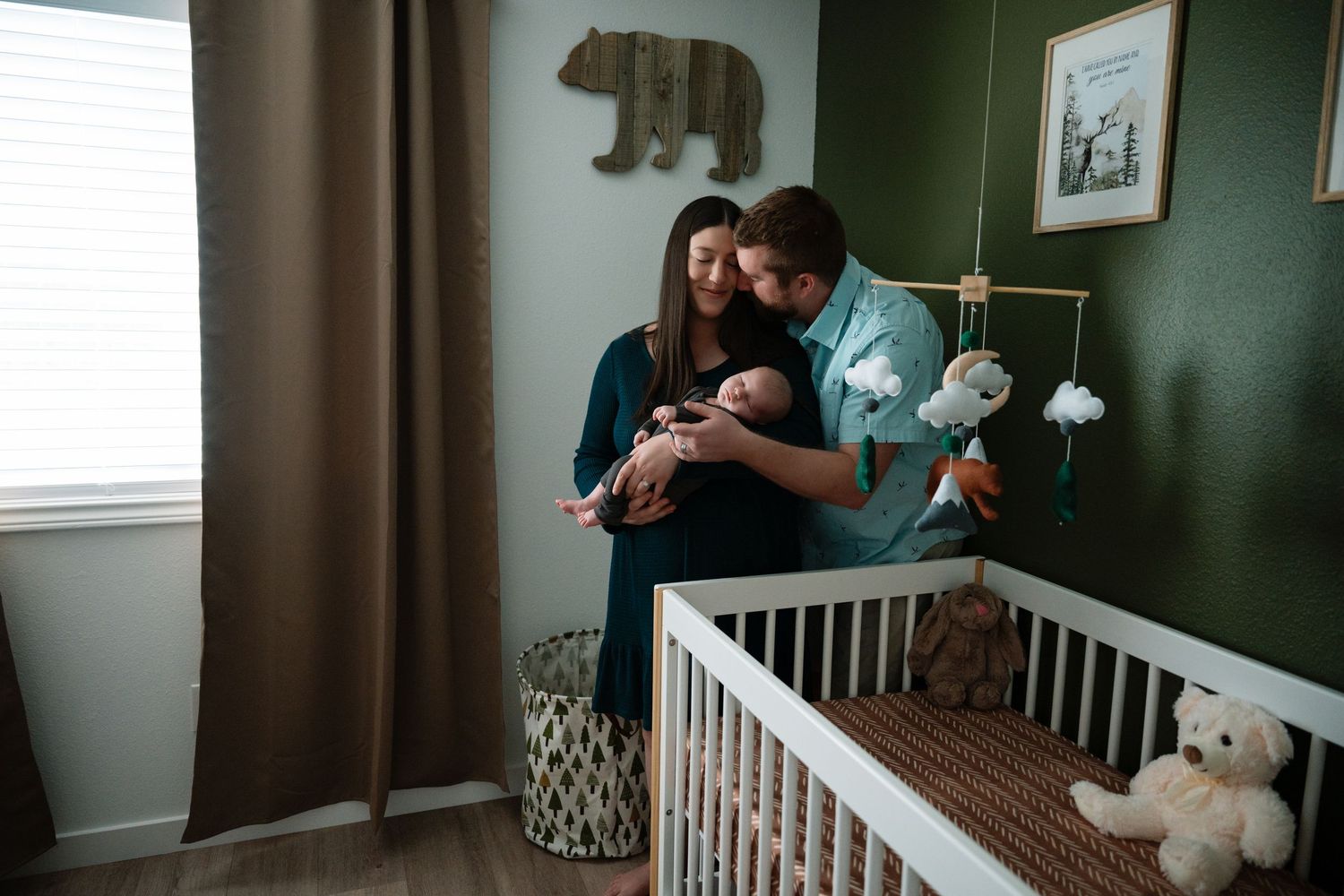 New parents tenderly hold their newborn baby in a green nursery with a white crib and bear decor.