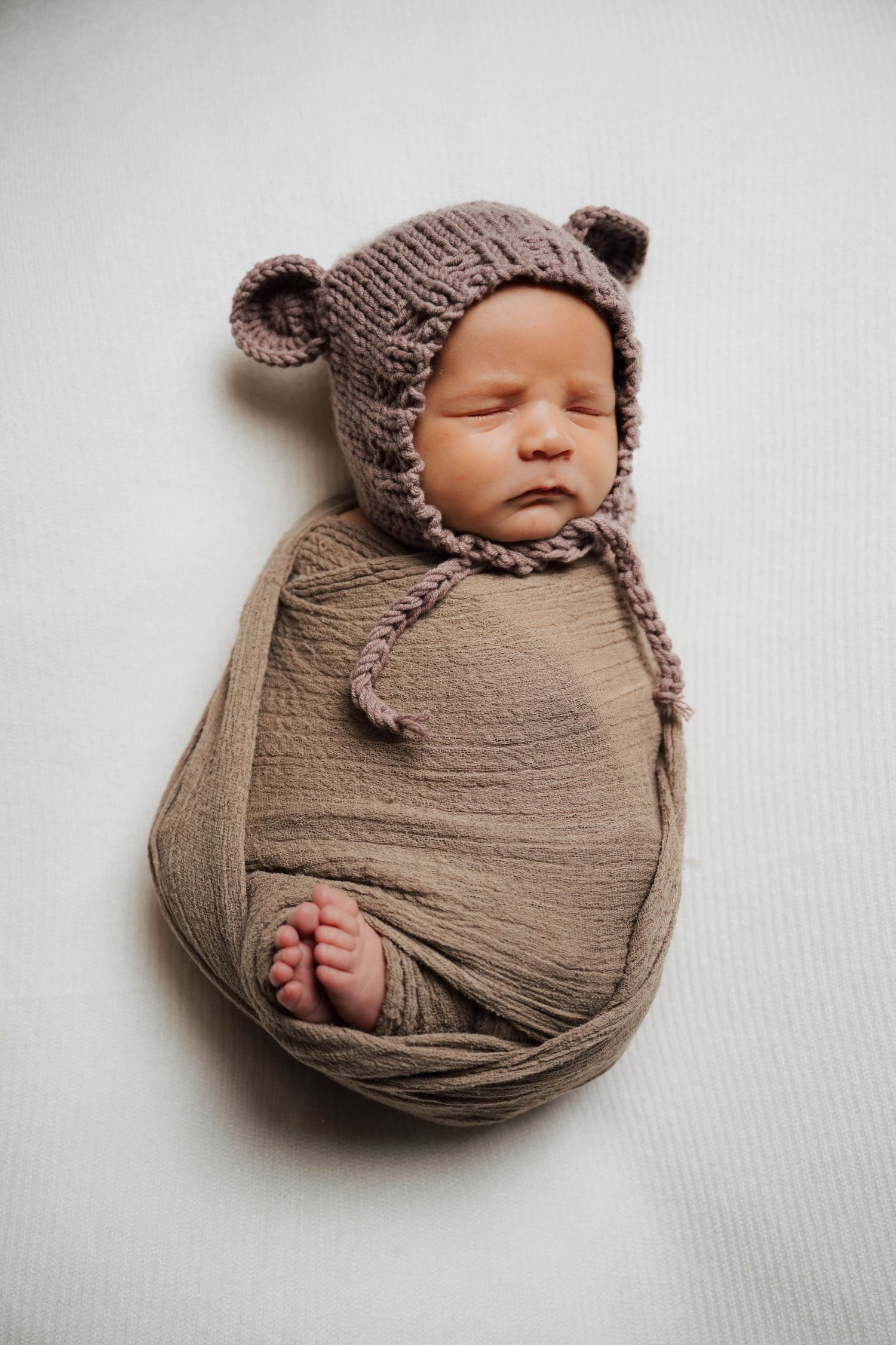 Newborn baby swaddled in brown wrap wearing a knitted bear bonnet hat, sleeping peacefully on white background.