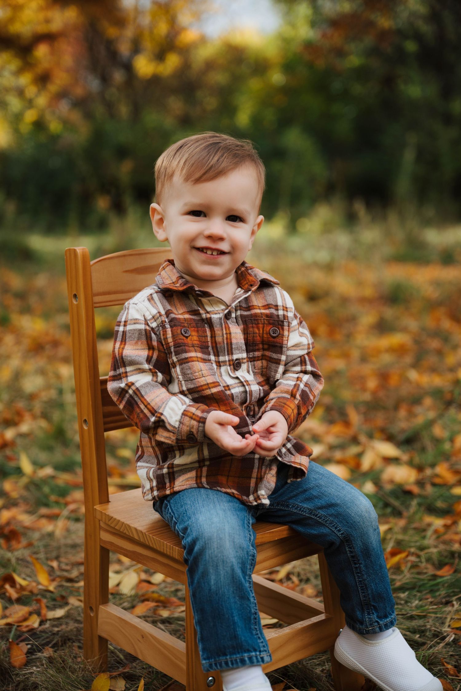 A small child sits in a wooden chair outdoors among fallen autumn leaves wearing a plaid shirt and jeans.