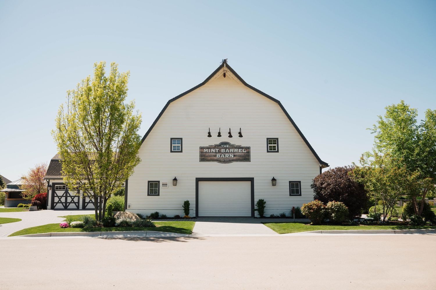 White barn wedding venue with gambrel roof, large garage door, and lush green trees under a clear blue sky.