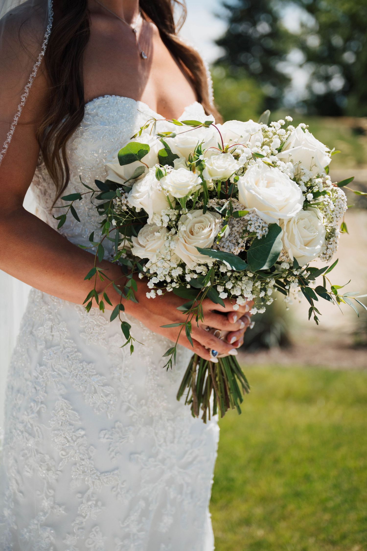 Bride in white lace gown holding elegant bridal bouquet of white roses and greenery on wedding day.