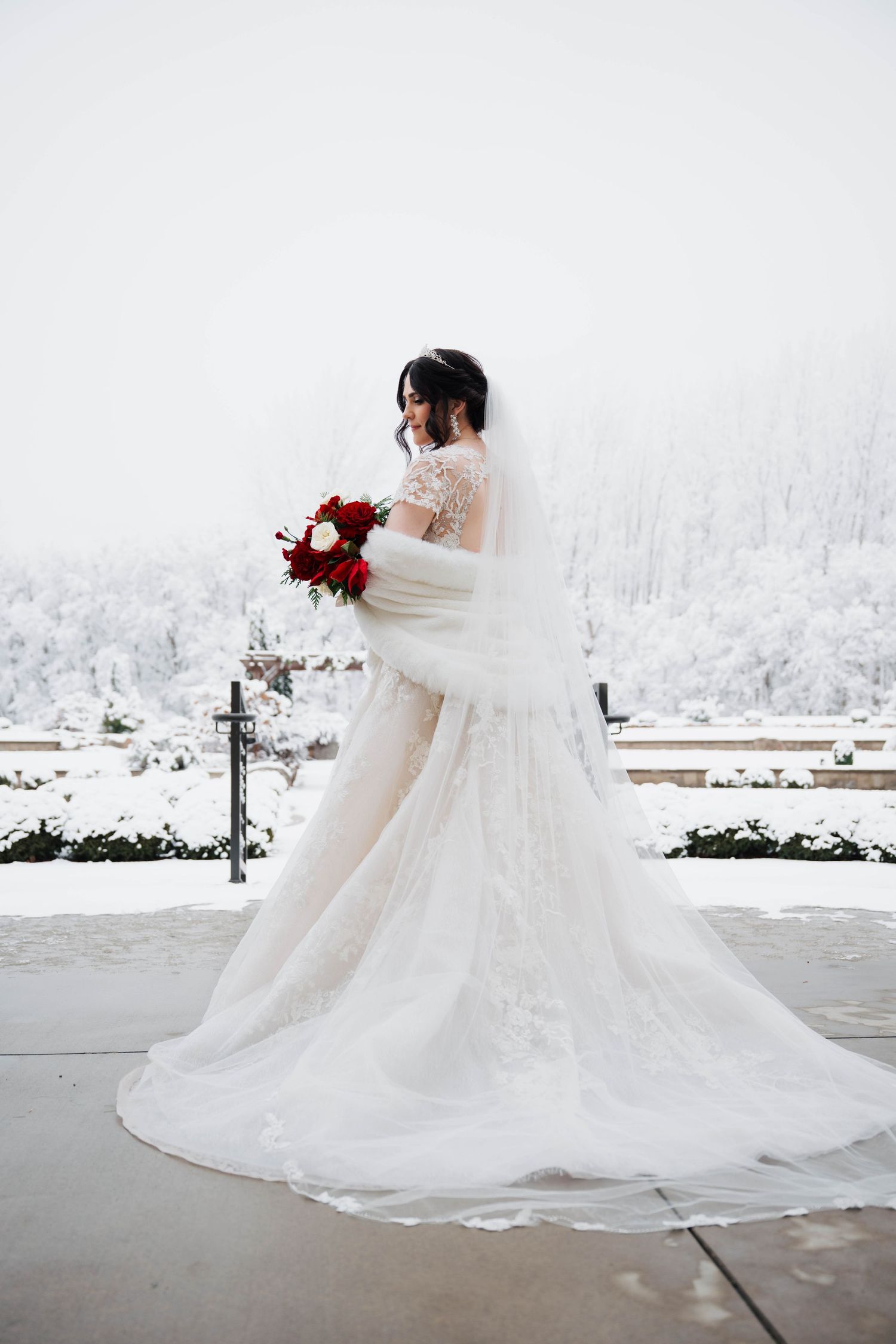 Bride in white lace gown with veil holds red bouquet outdoors in snowy winter landscape.