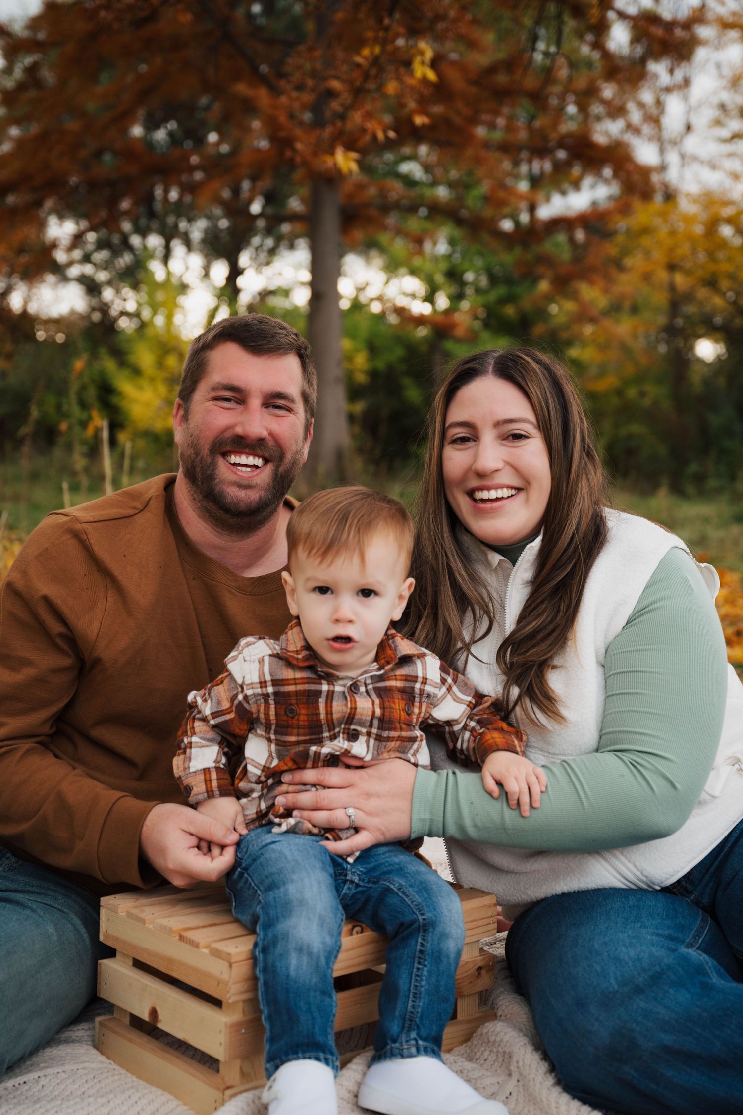 A family sits together on wooden pallets during an autumn outdoor photo session with warm fall colors in the background.