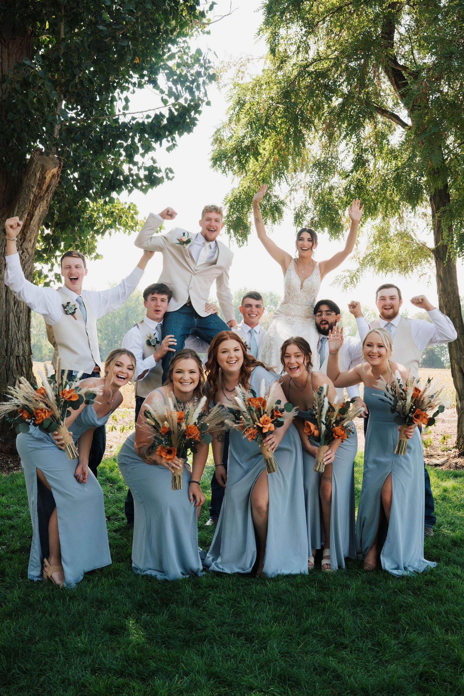 Wedding party celebrating outdoors, bridesmaids in blue gowns with bouquets, groomsmen in white, bride and groom elevated above group.