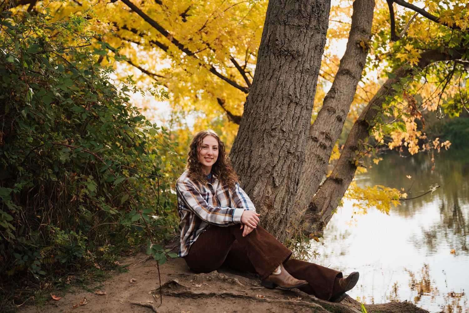 Person sitting by tree trunk with autumn leaves and water in background.