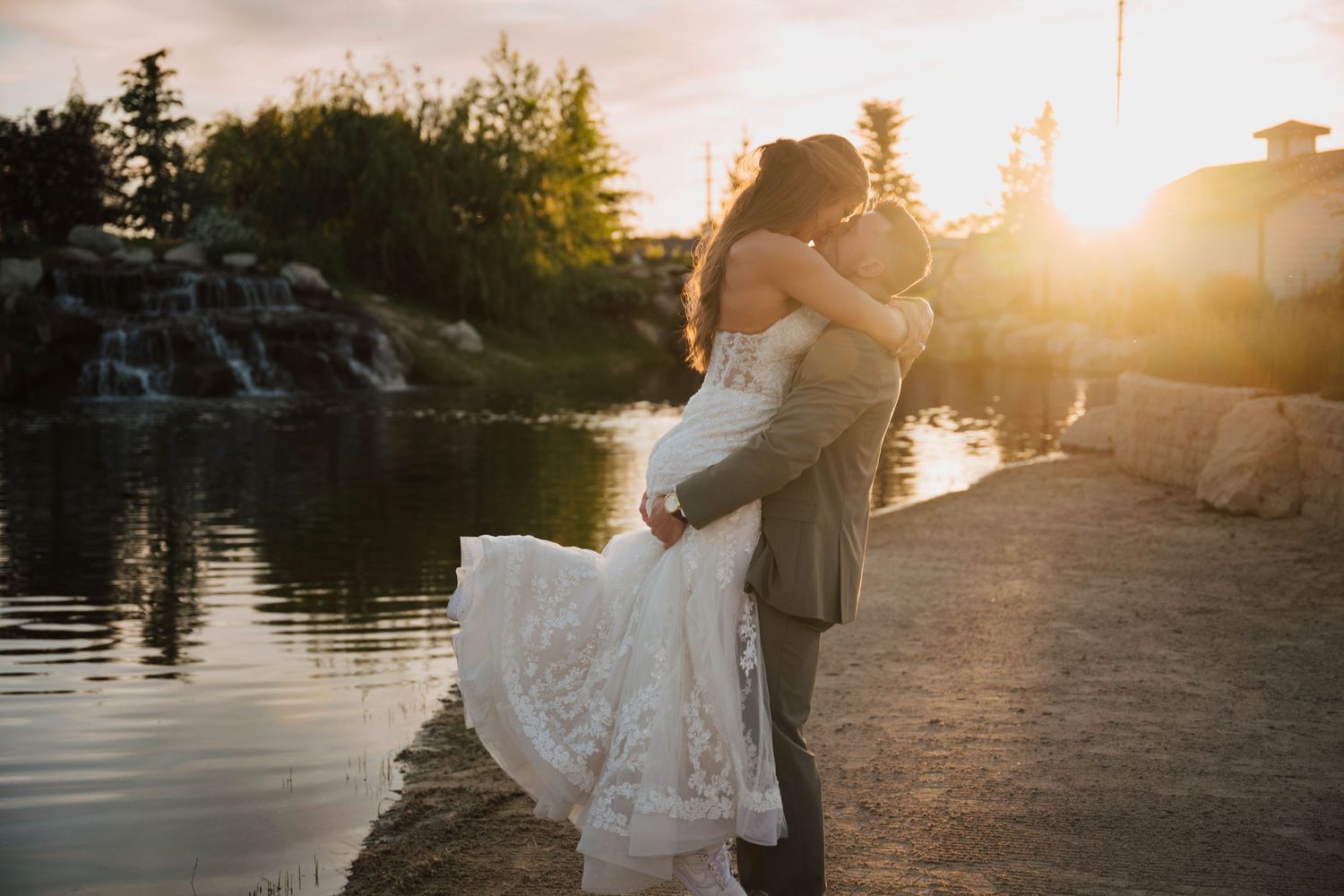 Couple sharing a romantic kiss at sunset by a serene pond, bride in lace gown, groom in gray suit.