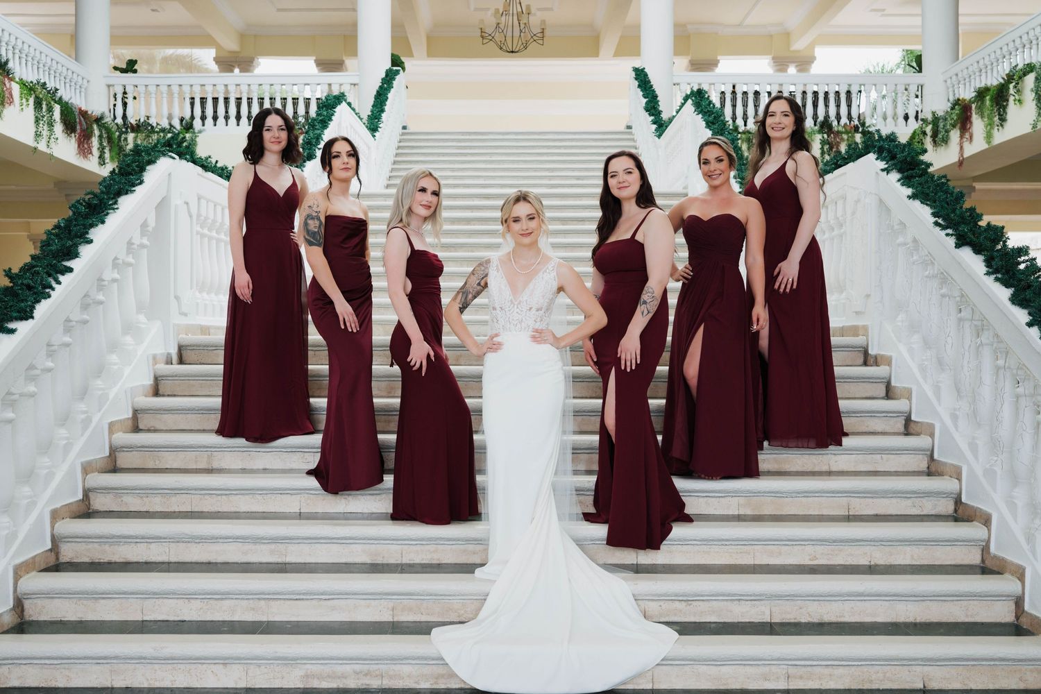 Bride in white gown poses with six bridesmaids in burgundy dresses on elegant white marble staircase.