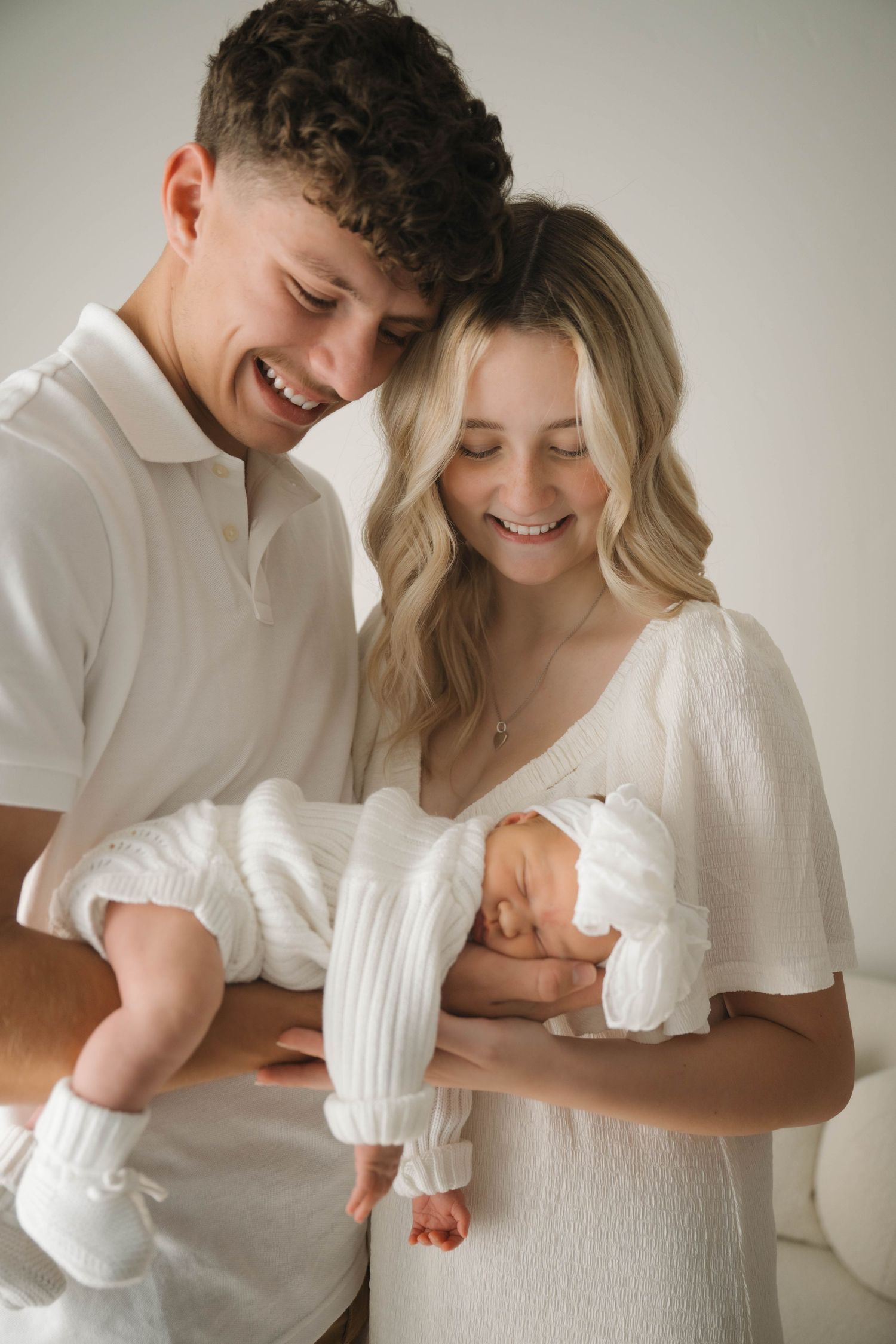 Happy young couple in white outfits smiling while cradling their newborn baby wrapped in white.