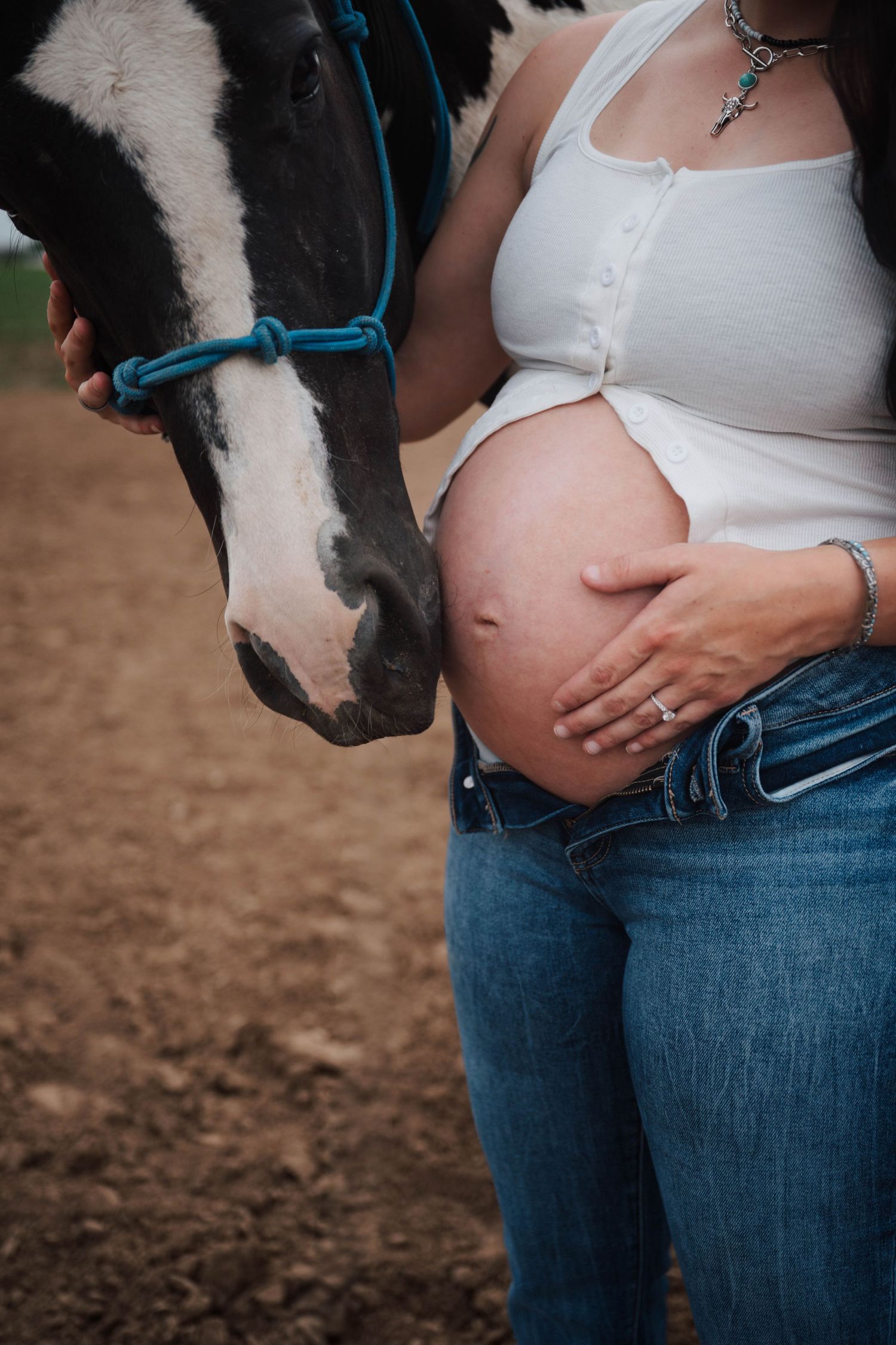 Pregnant woman in white tank top and jeans holds her bare belly next to a black and white horse with blue halter.