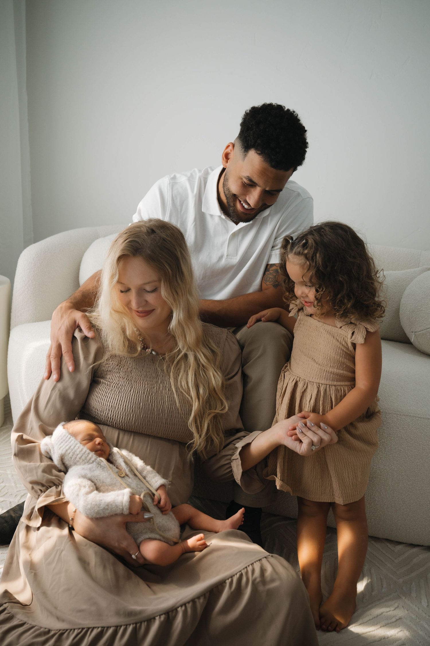 A happy interracial family sits together on a couch, admiring their newborn baby with a young daughter nearby.