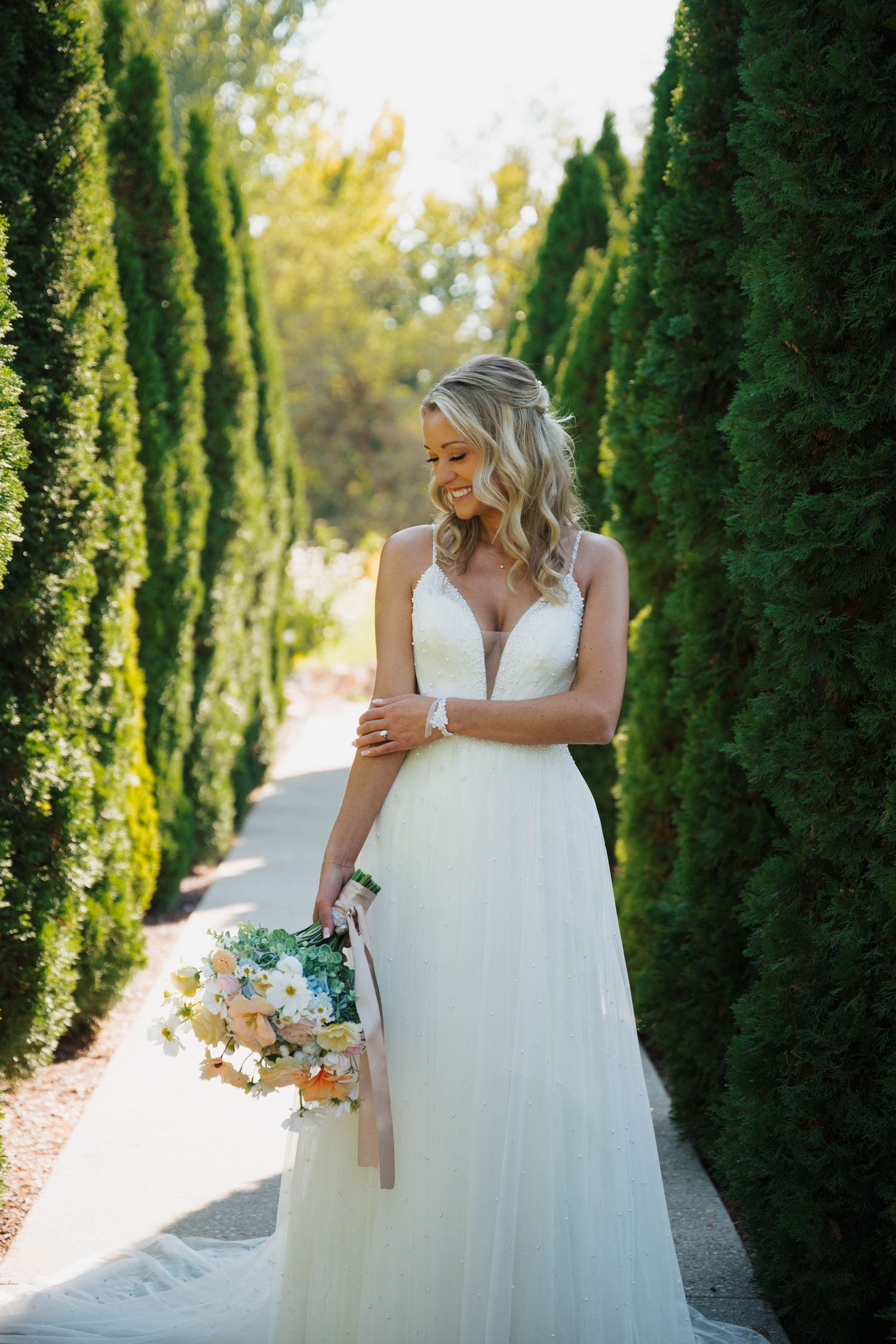 Bride in white V-neck gown holds floral bouquet while standing on a sunlit path lined with tall cypress trees.