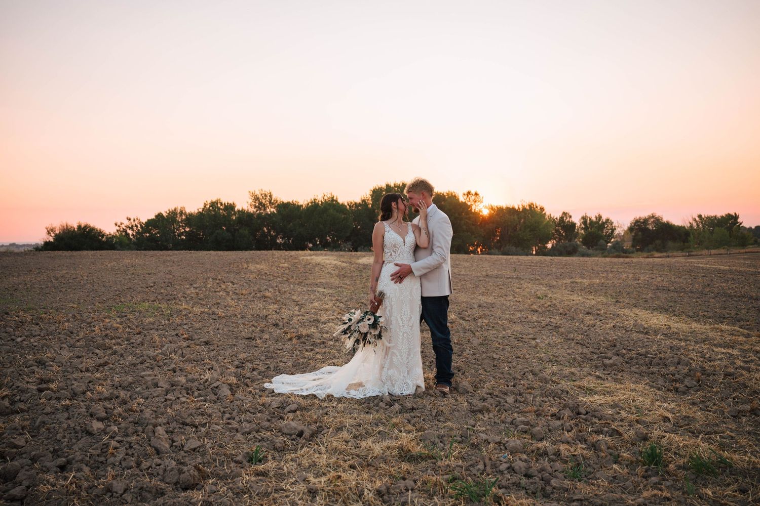 Bride and groom share a kiss at sunset in an open field, bride holding a bouquet in a lace wedding gown.