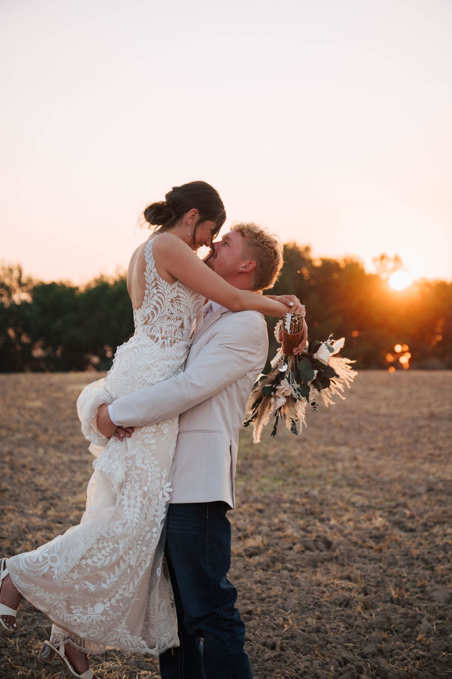 Groom lifting bride in lace dress at golden sunset in open field, sharing romantic kiss with bouquet.