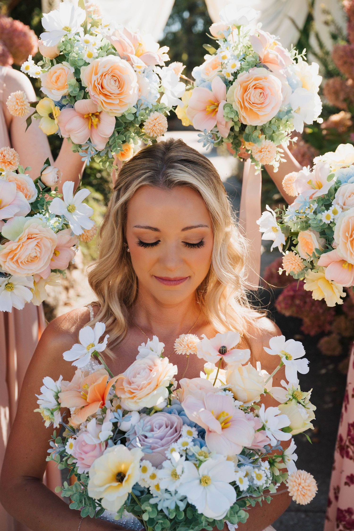 Bride with wavy blonde hair holds a pastel bouquet of roses and wildflowers, surrounded by peach and white floral arrangements.