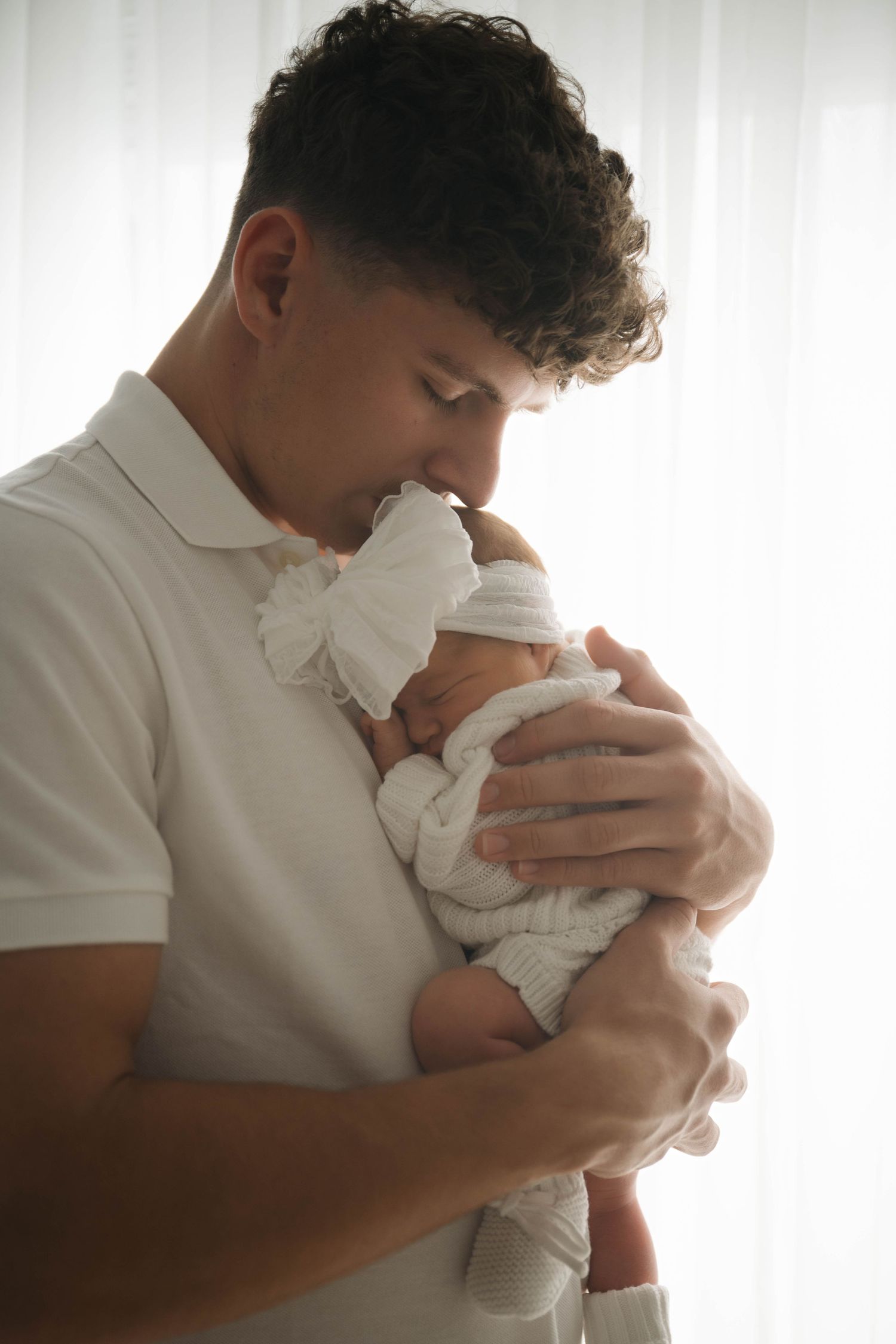 A father tenderly kisses his newborn baby wrapped in white, standing by a bright window.