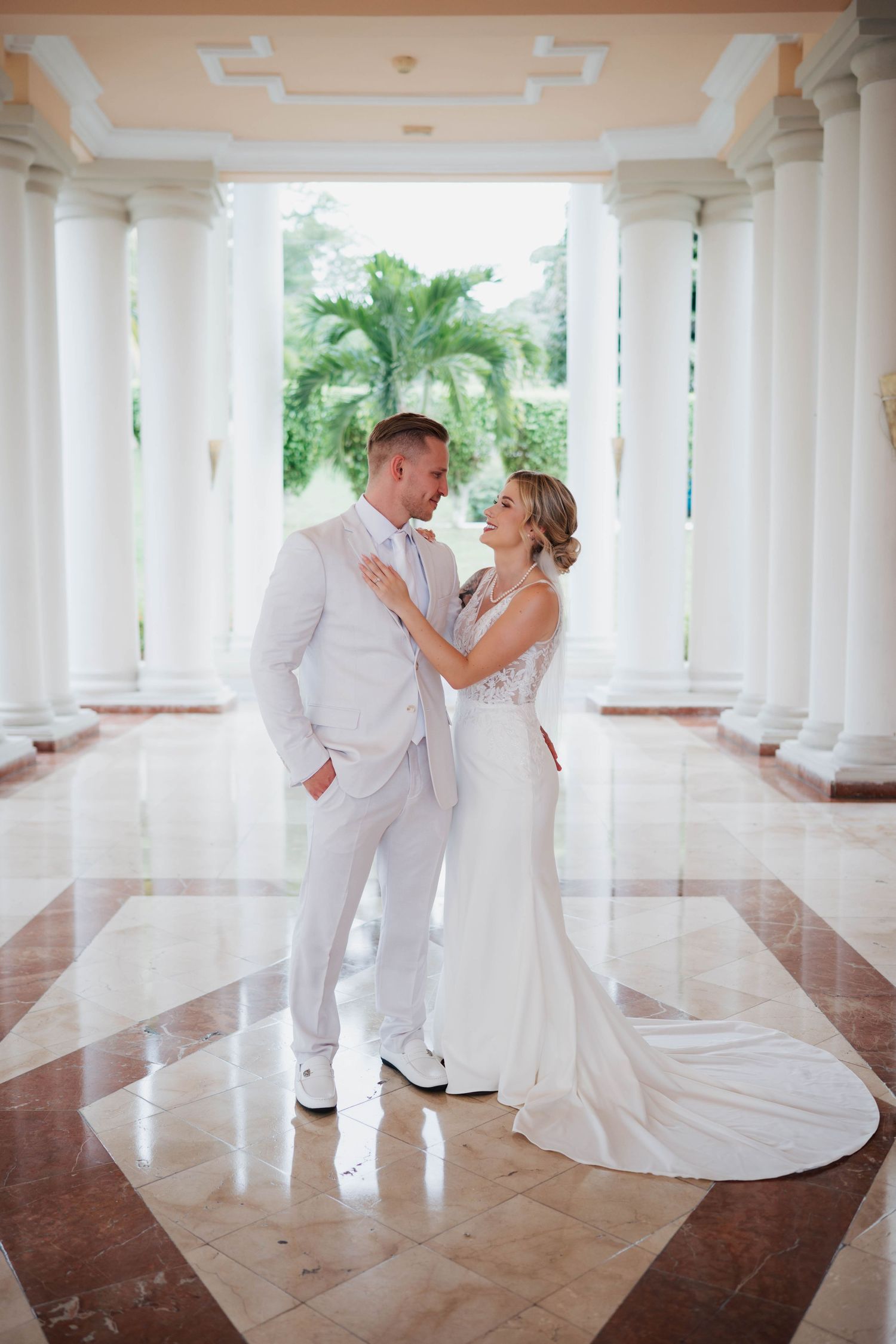 Bride and groom share a romantic moment in a white columned hall with marble floors and tropical greenery.