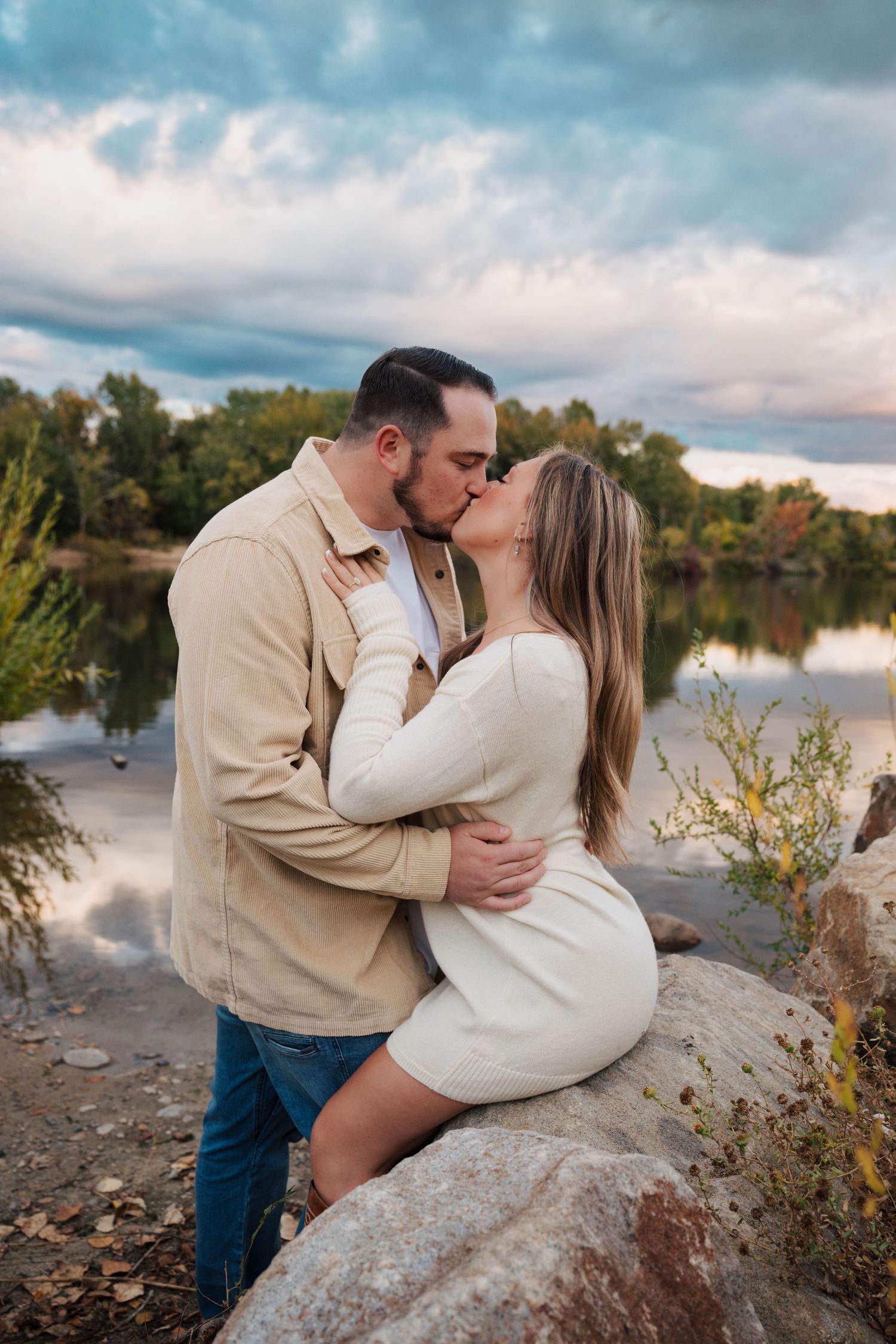 Romantic couple embracing on rocky shoreline during cloudy sunset.