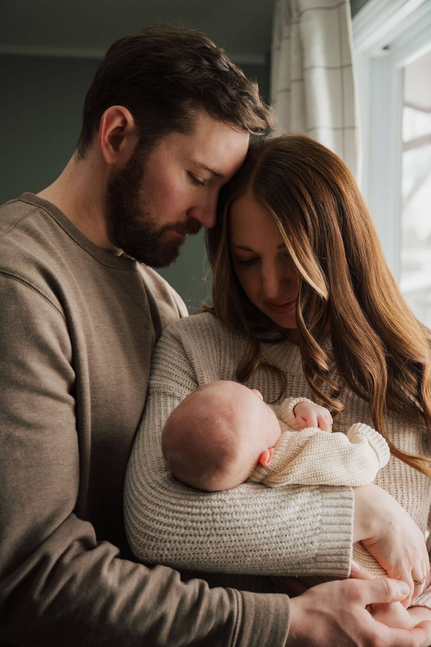 Tender family moment by window with parents holding newborn baby in knit sweaters.