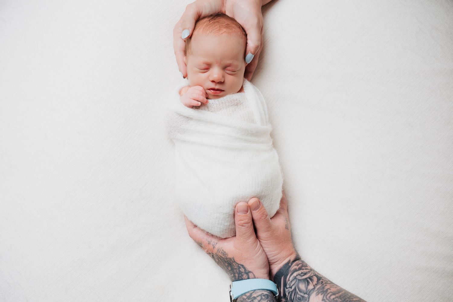 A sleeping newborn wrapped in white swaddle held gently by adult hands.