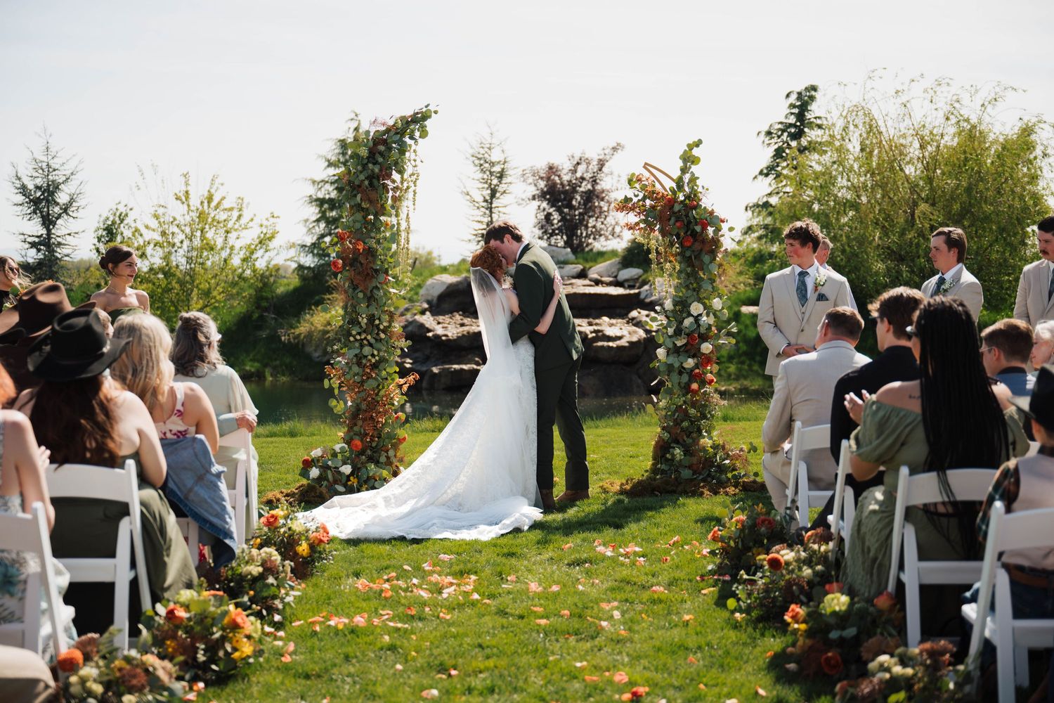 Outdoor wedding ceremony with couple kissing at floral arch, guests seated on white chairs, colorful blooms lining the aisle.