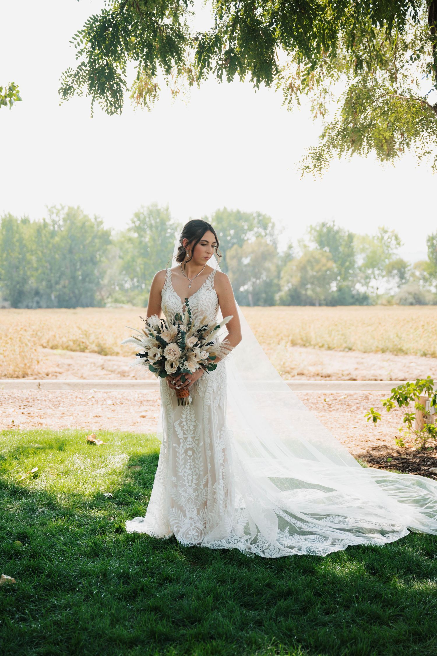 Bride in lace gown with long train holds white bouquet on green lawn with trees and field backdrop.