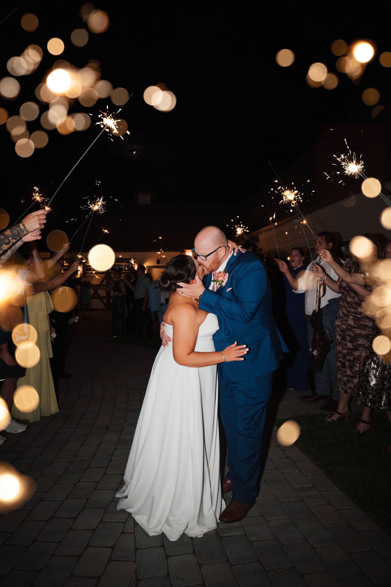 Newlyweds share a romantic kiss during a sparkler send-off at their outdoor wedding reception at night.
